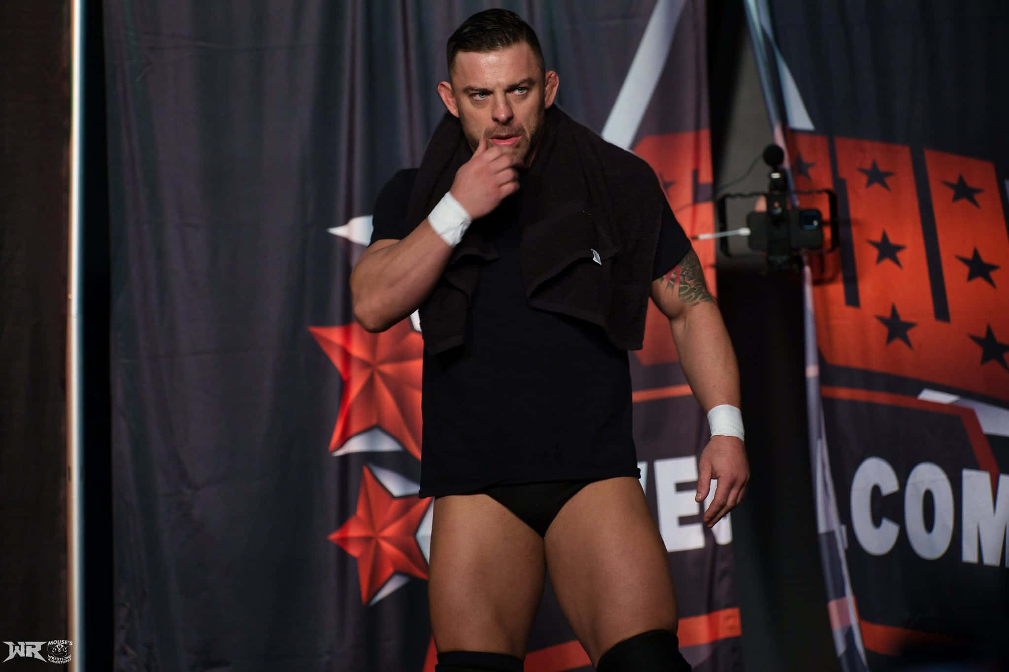 WWE 桌布 壁紙 — 一位肌肉發達的摔角手穿著黑色裝備，自信地站在舞台上，肩上搭著毛巾，背景是星星圖案。