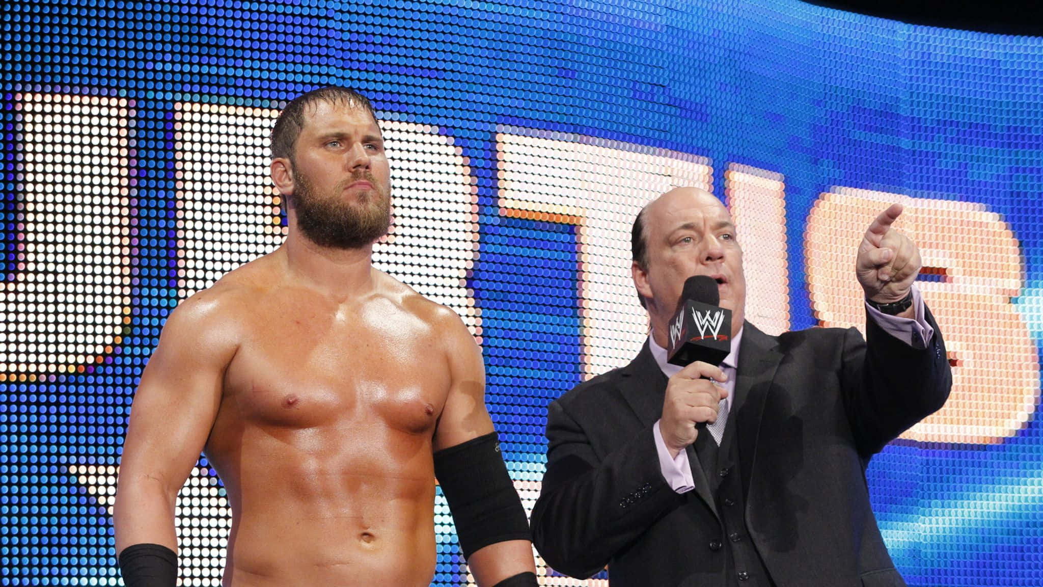 WWE 桌布 壁紙 — 一位肌肉發達的摔角手站在身穿西裝、手持WWE麥克風的主持人身旁，背景是明亮的藍色LED螢幕。