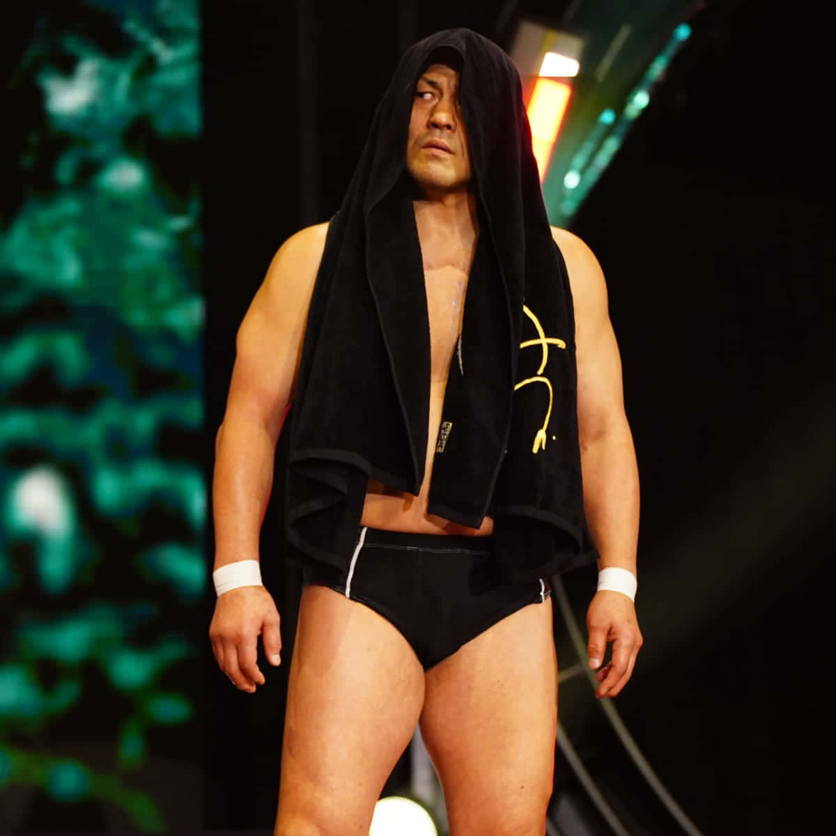 Aew 桌布 壁紙 — 一位肌肉發達的摔角手自信地站在舞台上，披著一條帶有黃色標誌的黑色毛巾，在戲劇性的綠色和橙色舞台燈光下。