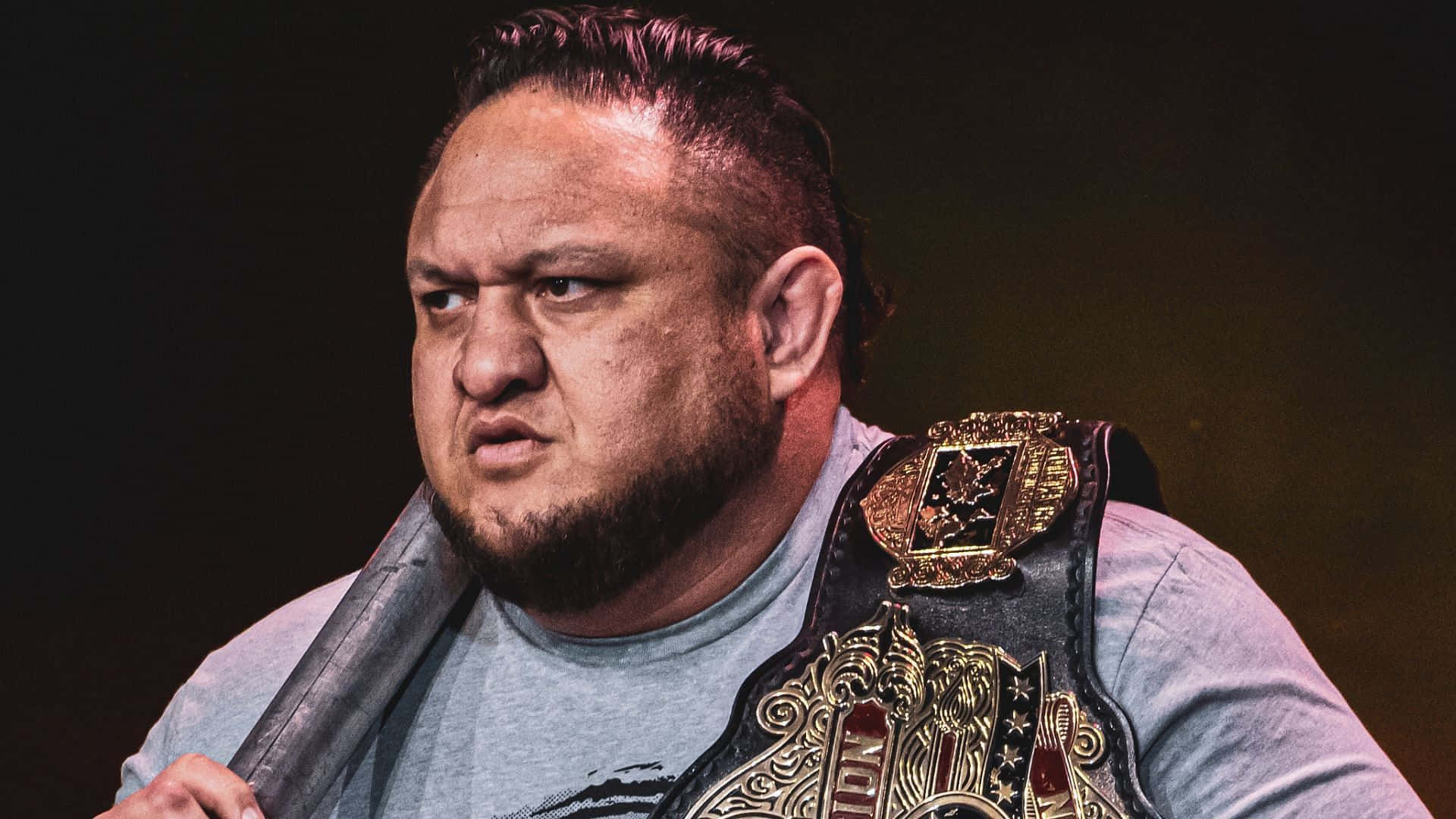 Aew 桌布 壁紙 — 一位肌肉發達的摔角手表情兇猛，身穿灰色T恤，肩上掛著冠軍腰帶，手持鋼椅。