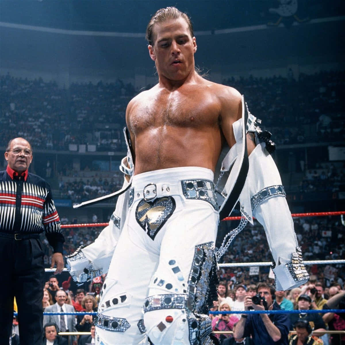shawn michaels 桌布 壁紙 — 一位肌肉發達的摔角手身穿白色亮片服裝，站在擂台上，裁判在他旁邊，觀眾在觀看。