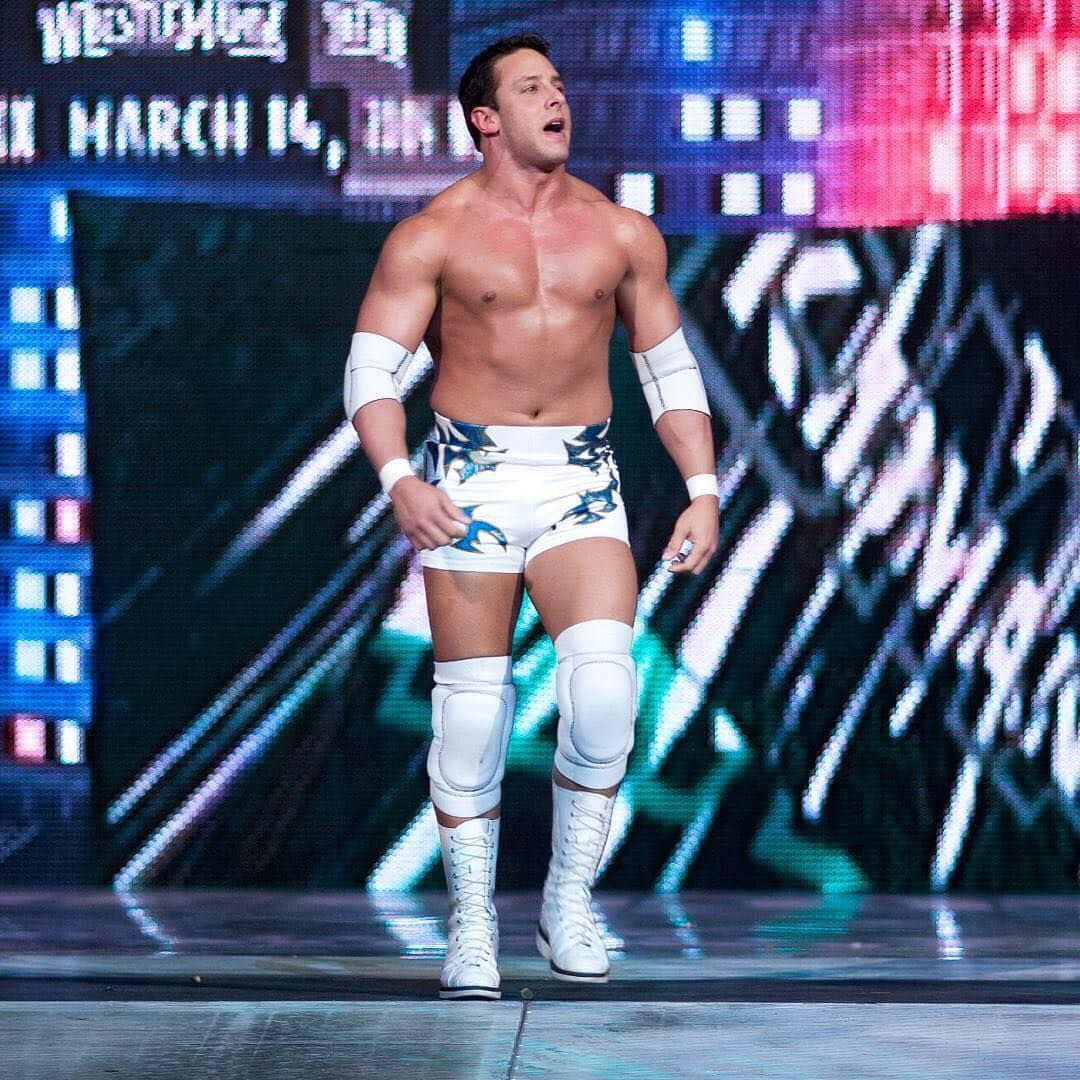 WWE 桌布 壁紙 — 一位肌肉發達的摔角手身穿白色裝備，自信地站在燈光璀璨的舞台上，背景是充滿活力的數位畫面。