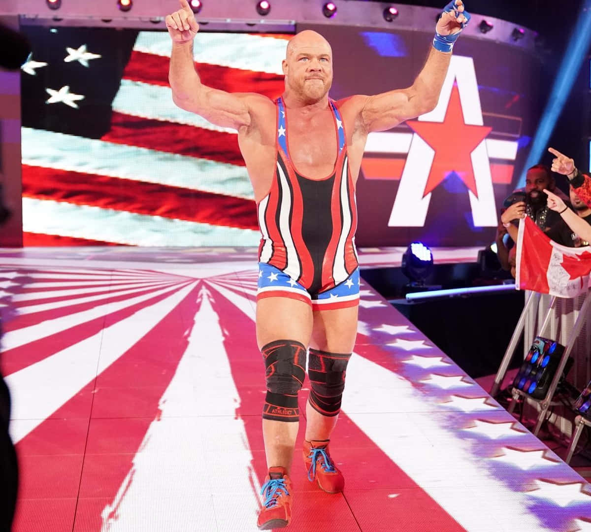 WWE 桌布 壁紙 — 一位肌肉發達的摔角手身穿紅、白、藍愛國服裝，在星條主題的擂台入口處勝利地舉起拳頭。