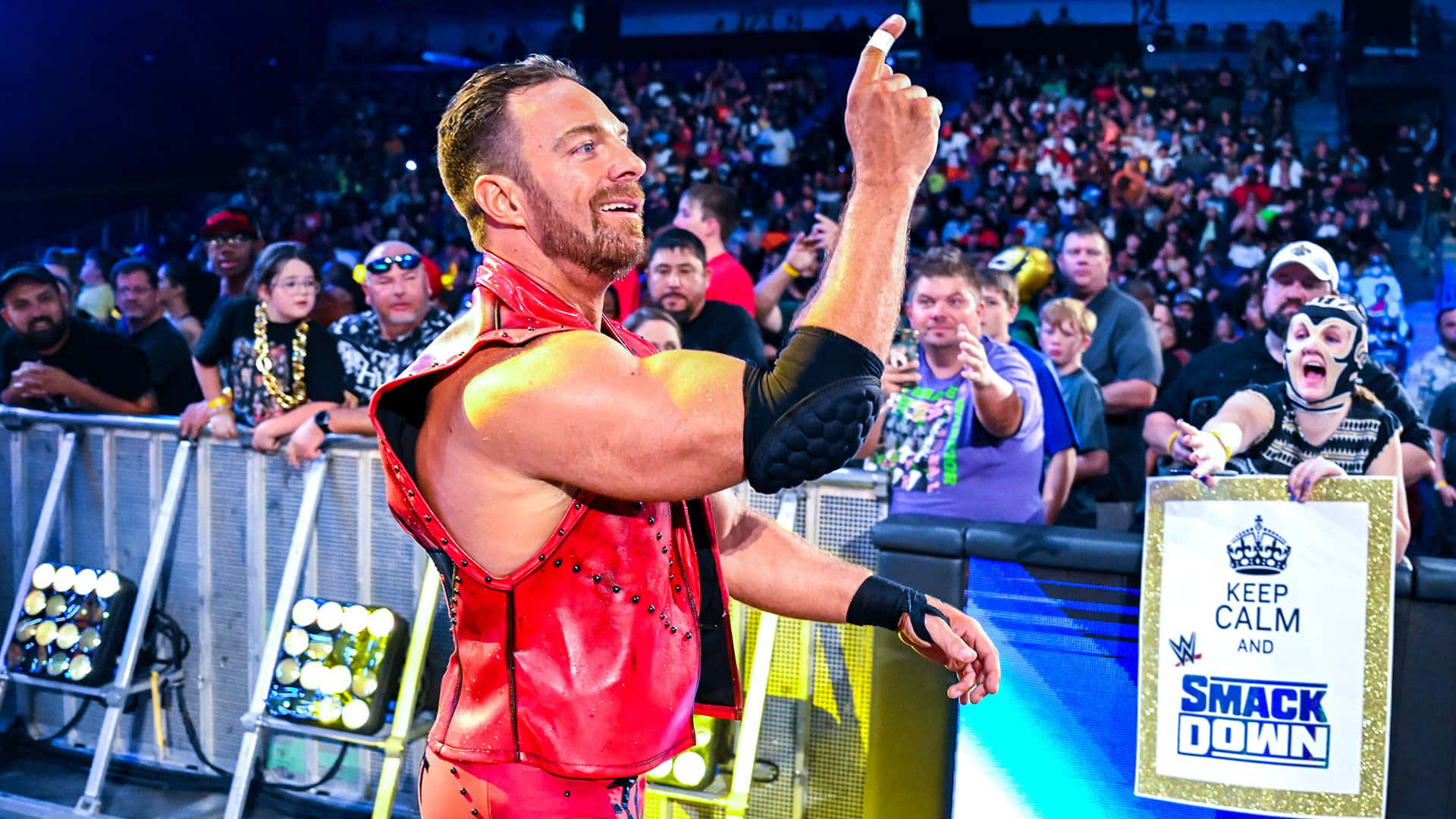WWE 桌布 壁紙 — 一位肌肉發達的摔角手身穿紅色皮革服裝，在燈光明亮的舞台上微笑著向上指，面前是歡呼的人群。