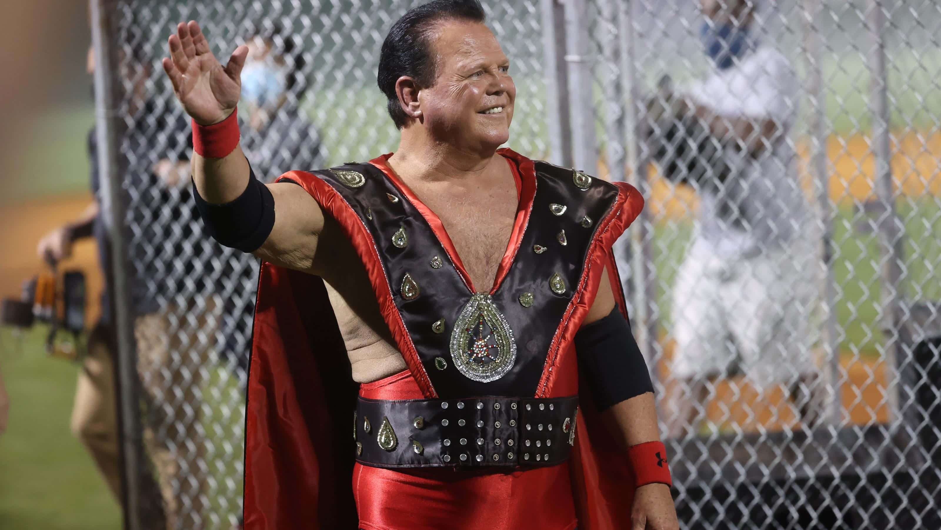 WWE 桌布 壁紙 — 一位肌肉發達的摔角手身穿紅黑服裝，在體育場燈光下隔著鐵絲網向粉絲揮手。
