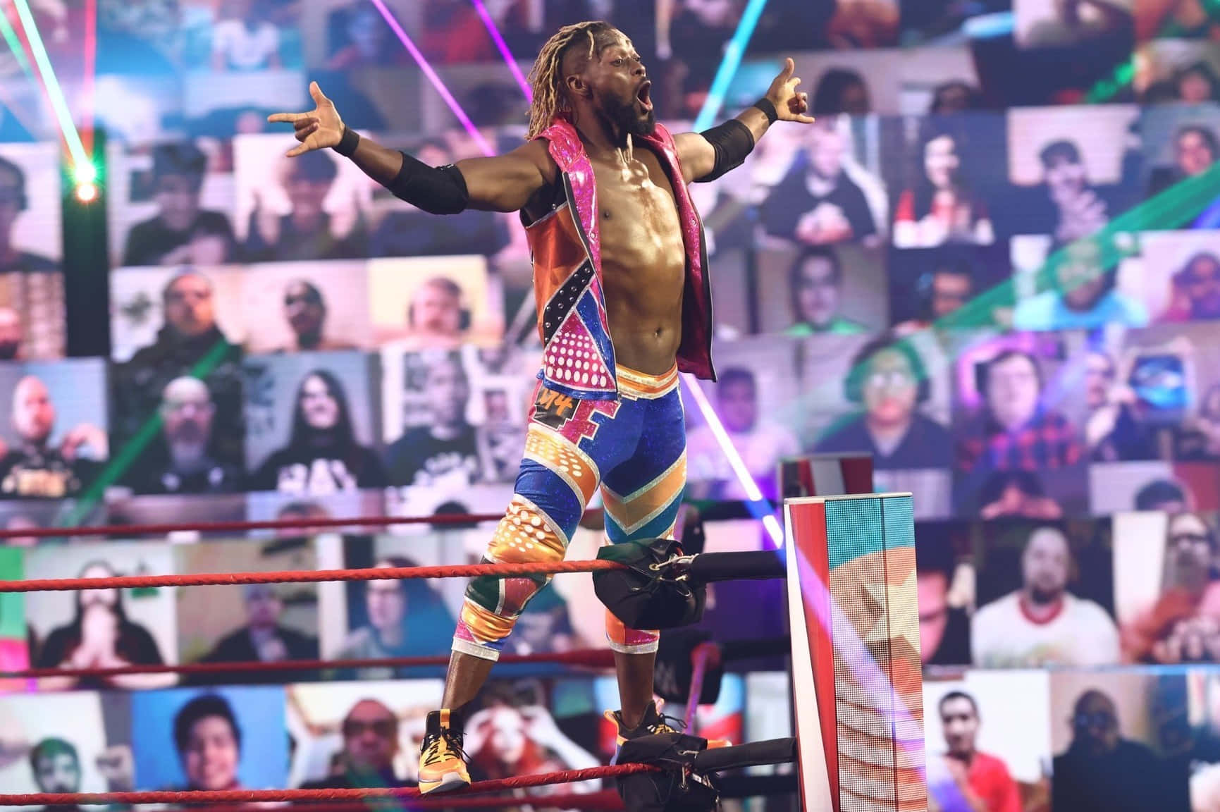 WWE 桌布 壁紙 — 一位肌肉發達的摔角手身穿色彩繽紛的裝備，站在摔角擂台的角柱上，雙臂張開，在明亮的舞台燈光下勝利地咆哮。