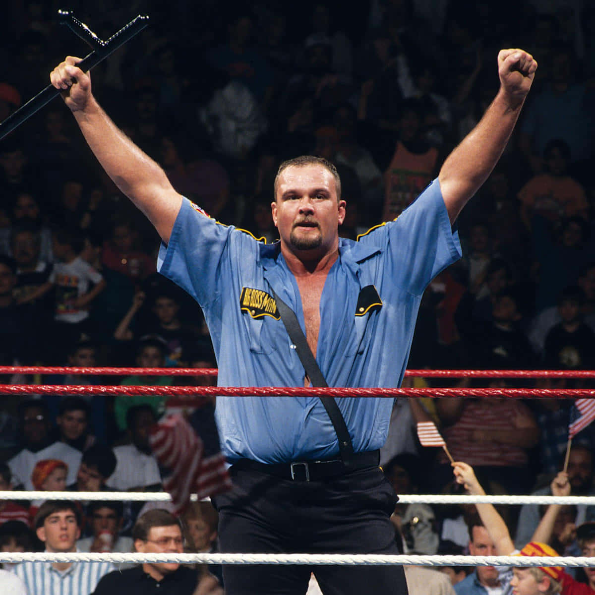 Big Boss Man 桌布 壁紙 — 一位肌肉發達的摔角手身穿藍色制服，在燈光明亮的擂台上，在歡呼的人群面前勝利地舉起拳頭和警棍。