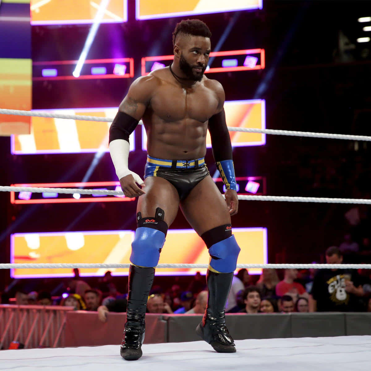 WWE 桌布 壁紙 — 一位肌肉發達的摔角手身穿藍黑裝備，自信地站在燈光明亮的擂台上，身後有發光的螢幕。