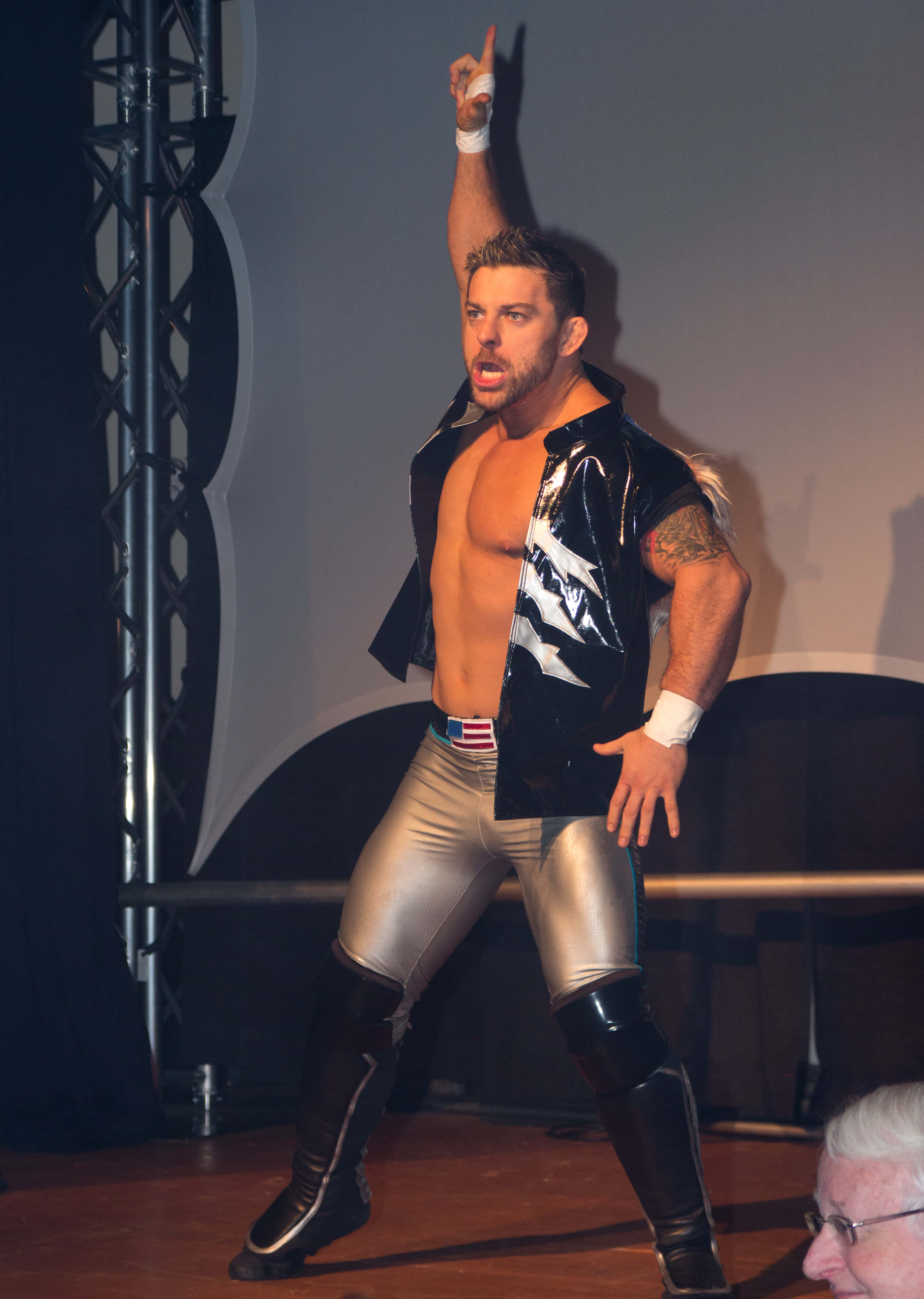 WWE 桌布 壁紙 — 一位肌肉發達的摔角手身穿閃亮的黑色和銀色裝備，在舞台上舉起一隻手臂擺出勝利姿勢。