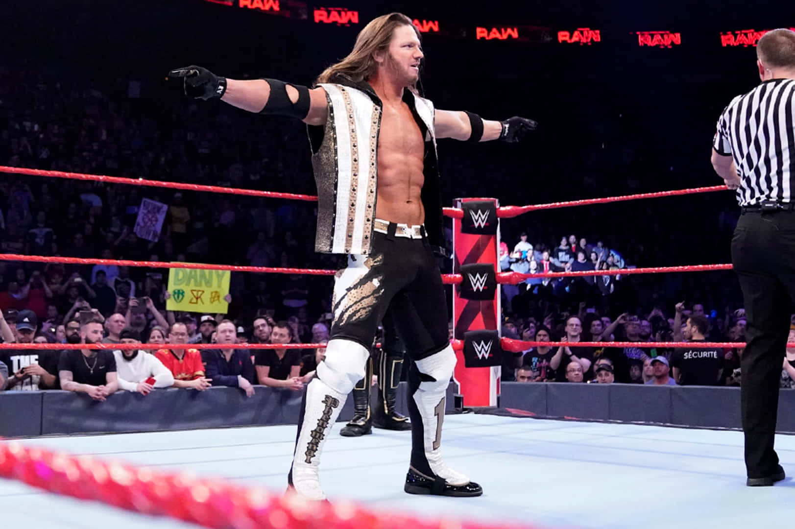 WWE 桌布 壁紙 — 一位肌肉發達的摔角手身穿黑白服裝，在摔角場上張開雙臂勝利地站立，裁判在旁觀看，背景中粉絲歡呼。