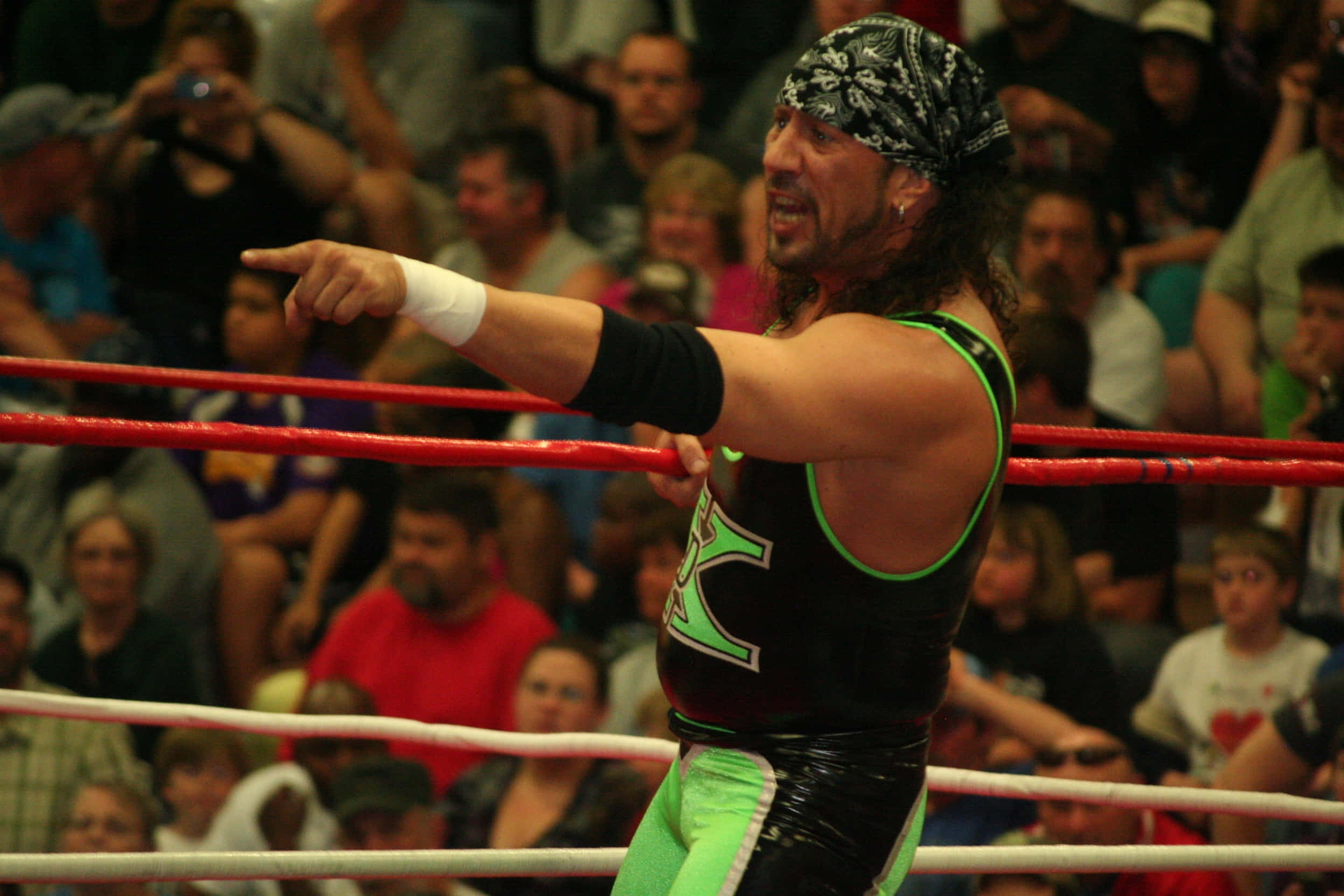 WWE 桌布 壁紙 — 一位肌肉發達的摔角手身穿黑色和螢光綠色裝備，在摔角場內戲劇性地指向人群。
