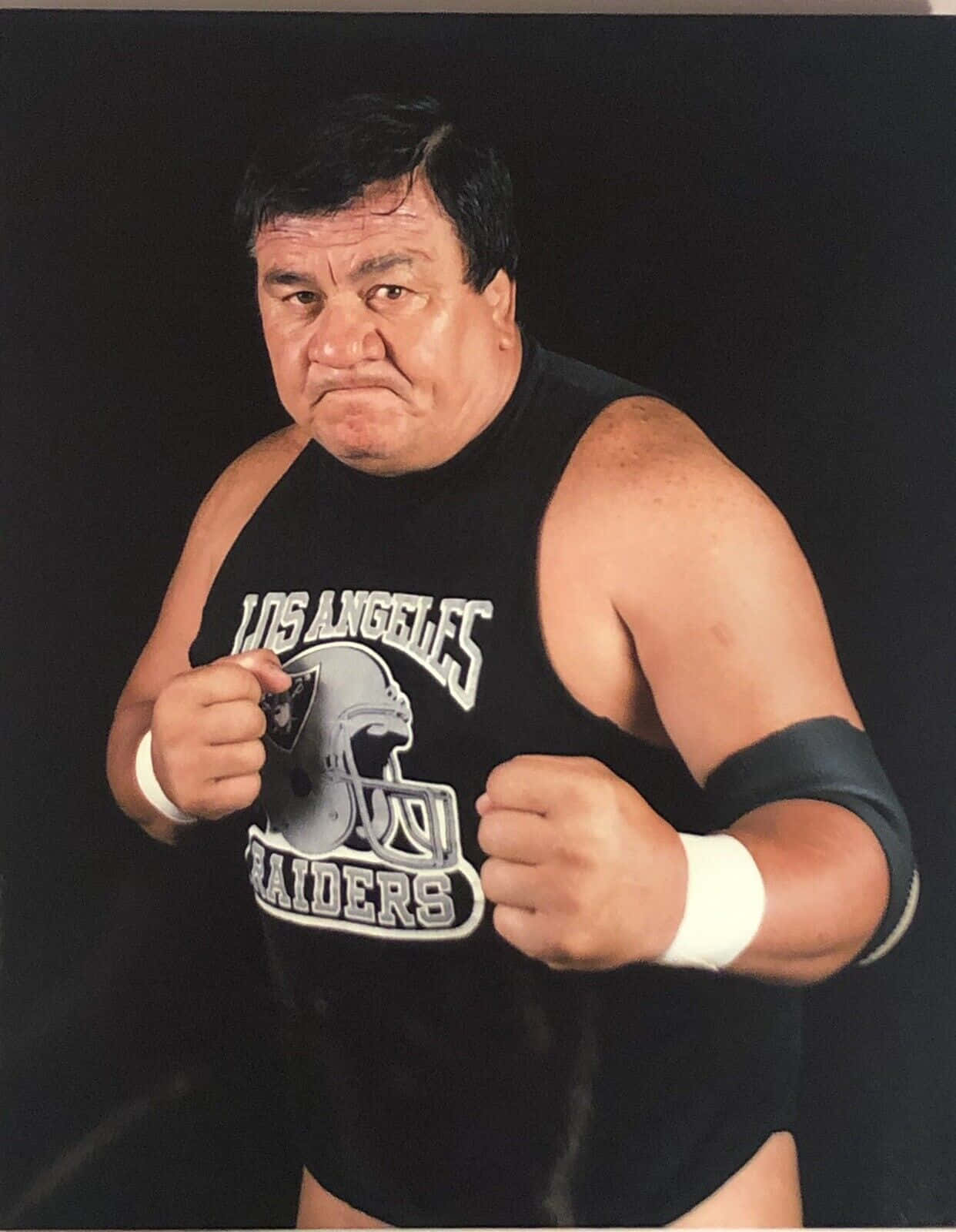 WWE 桌布 壁紙 — 一位肌肉發達的摔角手身穿黑色洛杉磯突襲者背心，在黑暗背景下握緊拳頭擺姿勢。