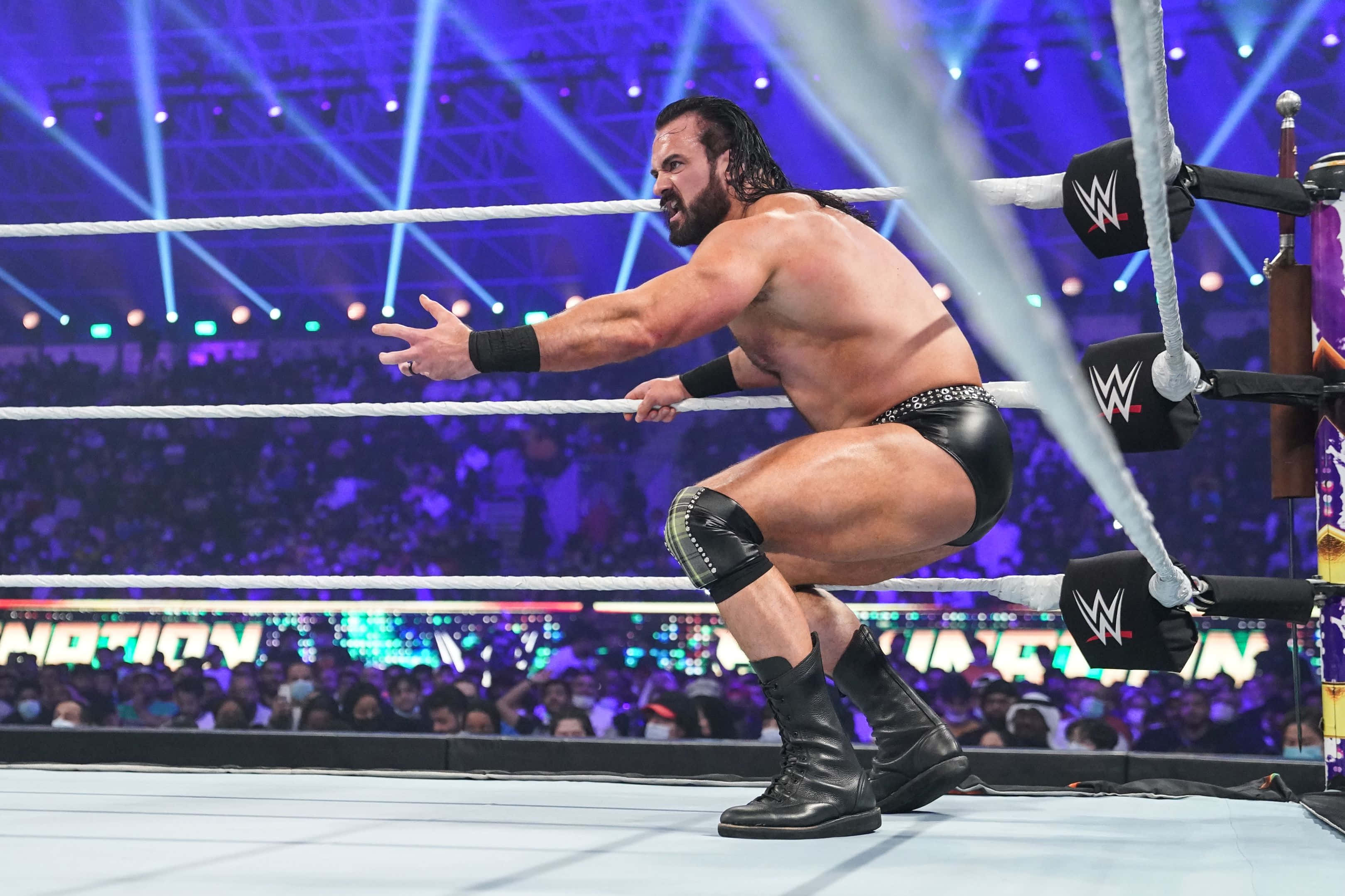 WWE 桌布 壁紙 — 一位肌肉發達的摔角手身穿黑色裝備，在明亮的藍色舞台燈光下蹲在摔角擂台上，身後是喧鬧的人群。