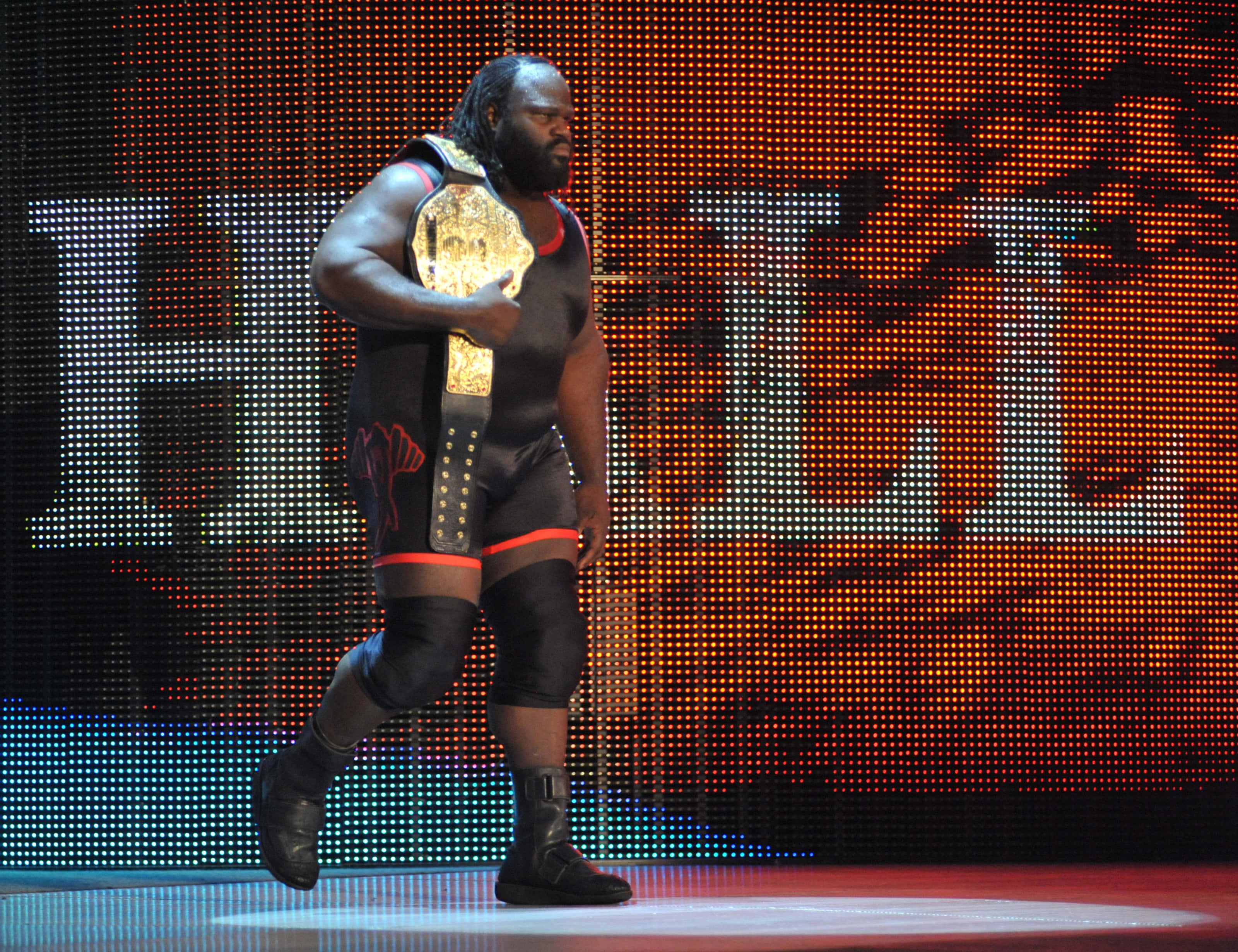 WWE 桌布 壁紙 — 一位肌肉發達的摔角手身穿黑色裝備，在明亮的場館燈光下自信地走上舞台，手持閃閃發光的冠軍腰帶。
