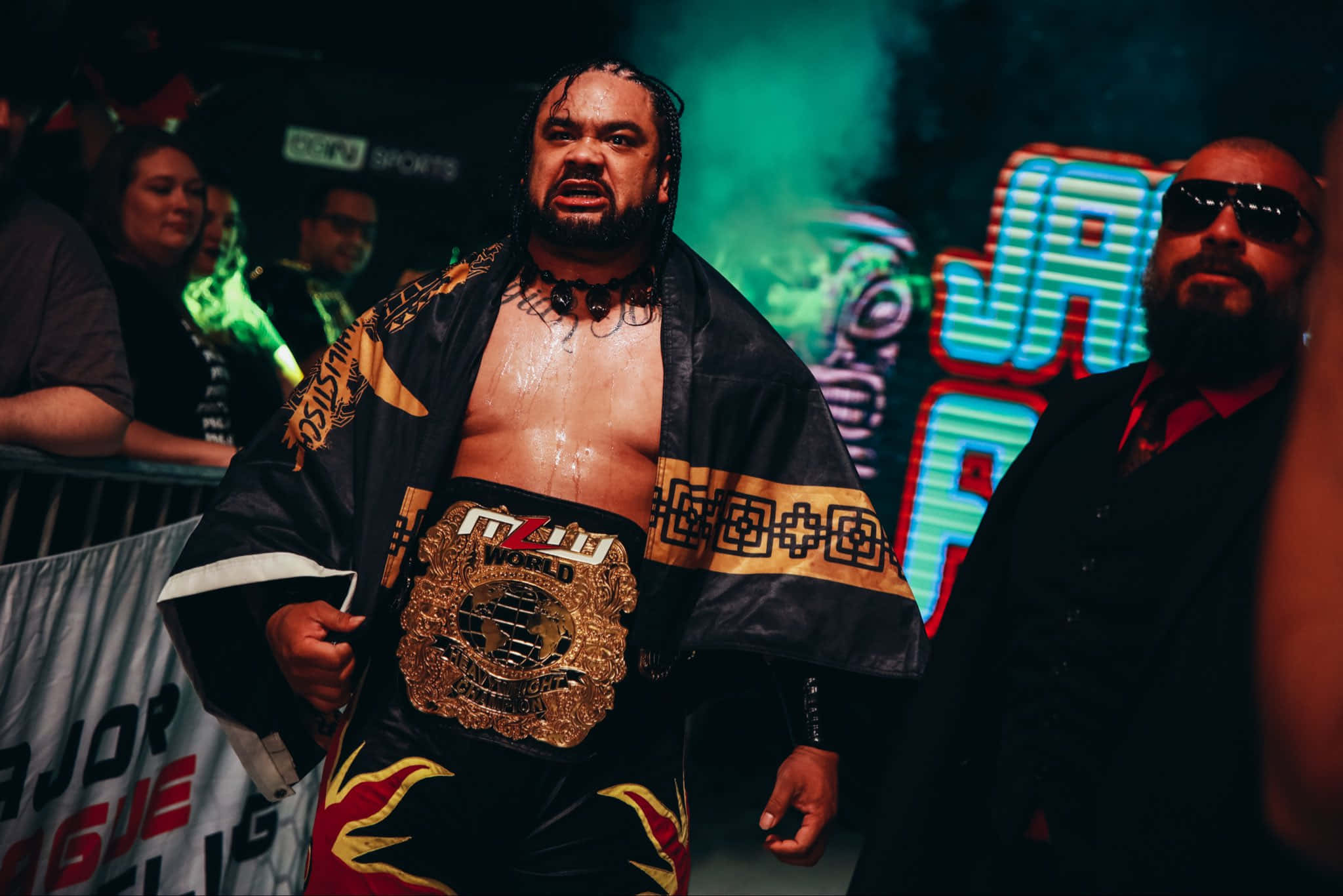WWE 桌布 壁紙 — 一位肌肉發達的摔角手身穿黑金色長袍，腰間繫著冠軍腰帶，在霓虹燈光下站在舞台中央，身後有群眾。