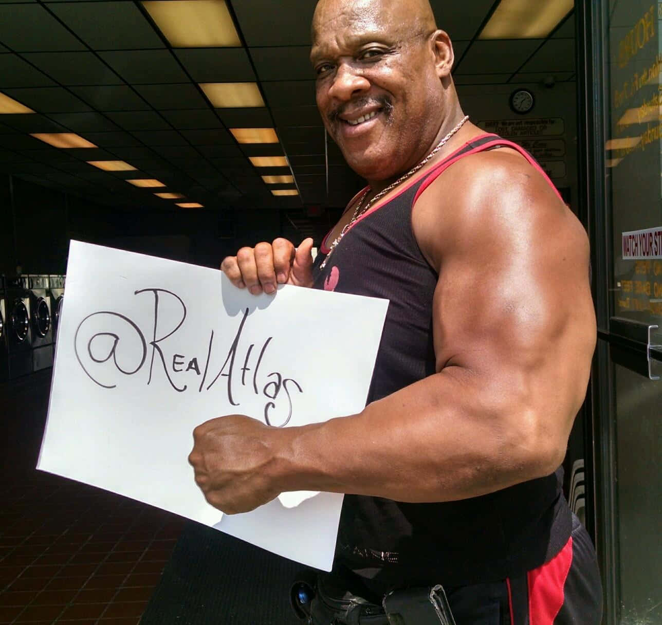 bodybuilding 桌布 壁紙 — 一位肌肉發達的男子在健身房微笑著展示肌肉，並舉著寫有 @RealAtlas 的牌子。