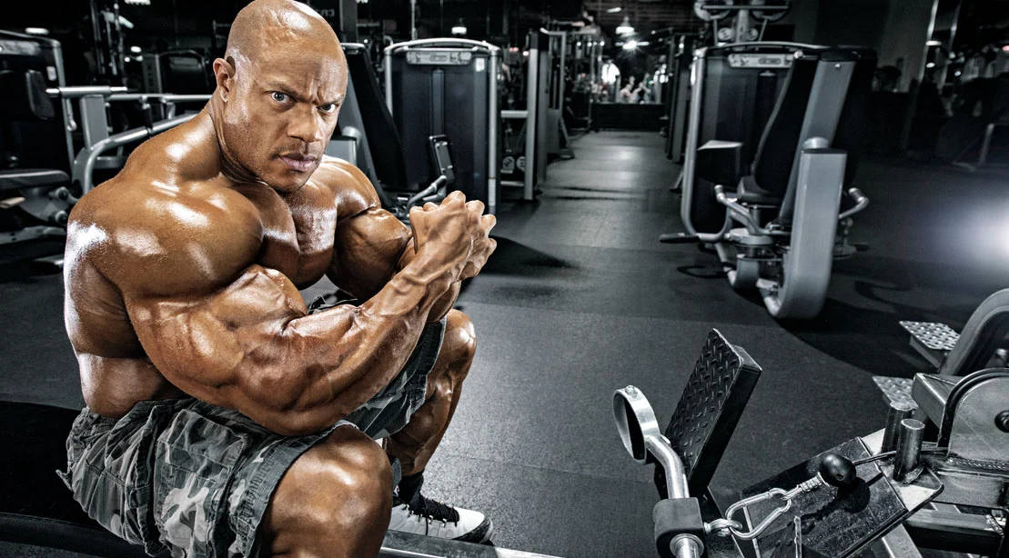 Exercise 桌布 壁紙 — 一位肌肉發達的男子坐在健身房的長椅上，周圍是重量訓練器材，他正彎曲手臂展示肌肉。
