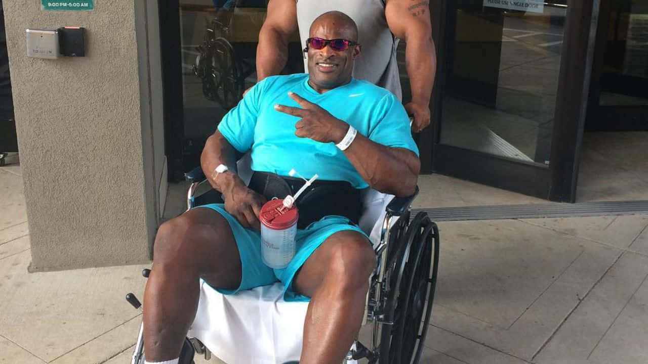 運動 桌布 壁紙 — 一位肌肉發達的男子坐在輪椅上，戴著太陽眼鏡和藍色襯衫，微笑著在建築物外拿著飲料。