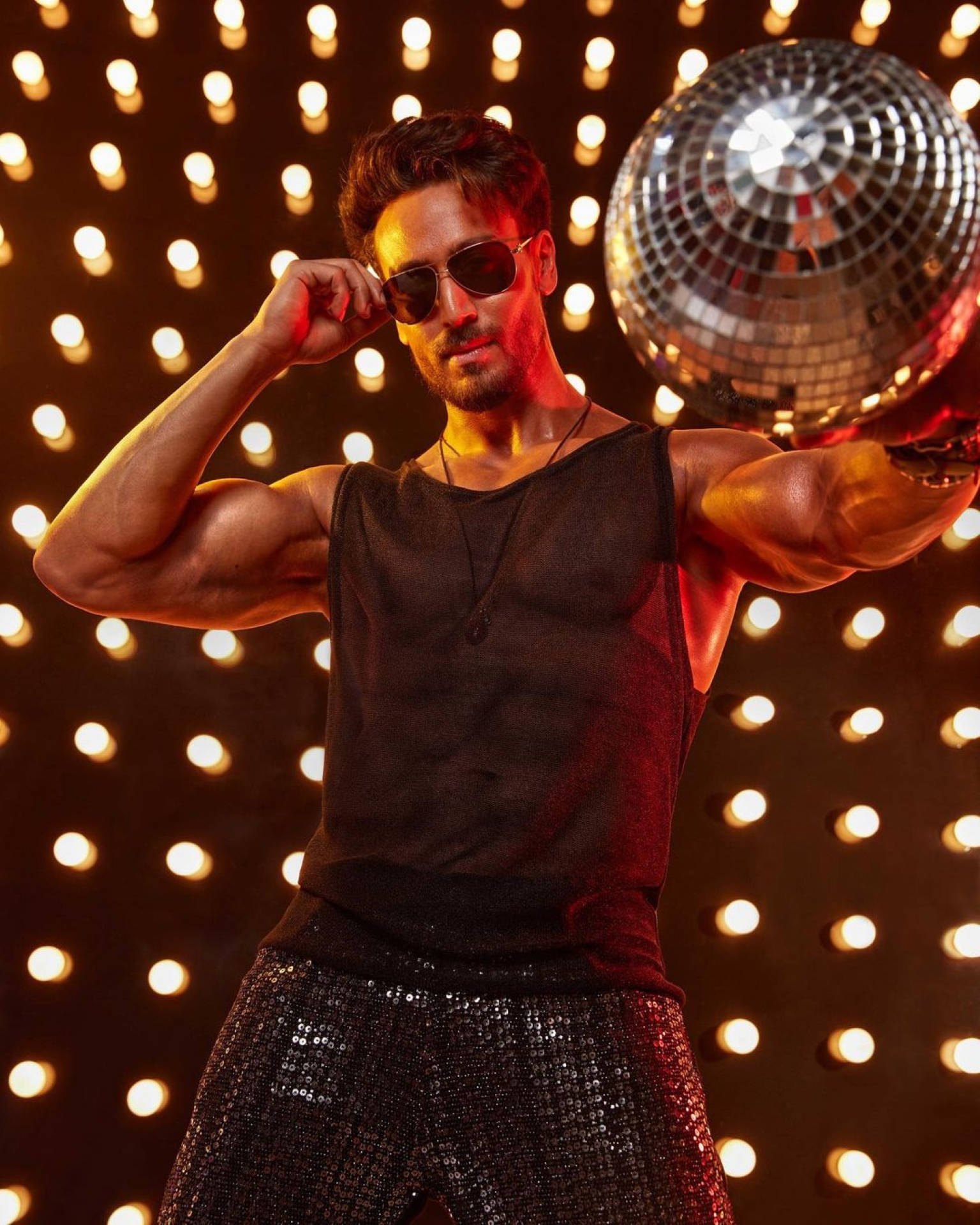 Tiger Shroff 桌布 壁紙 — 一位肌肉發達的男子穿著亮片褲和太陽眼鏡，在發光的燈牆旁自信地與迪斯科球合影。