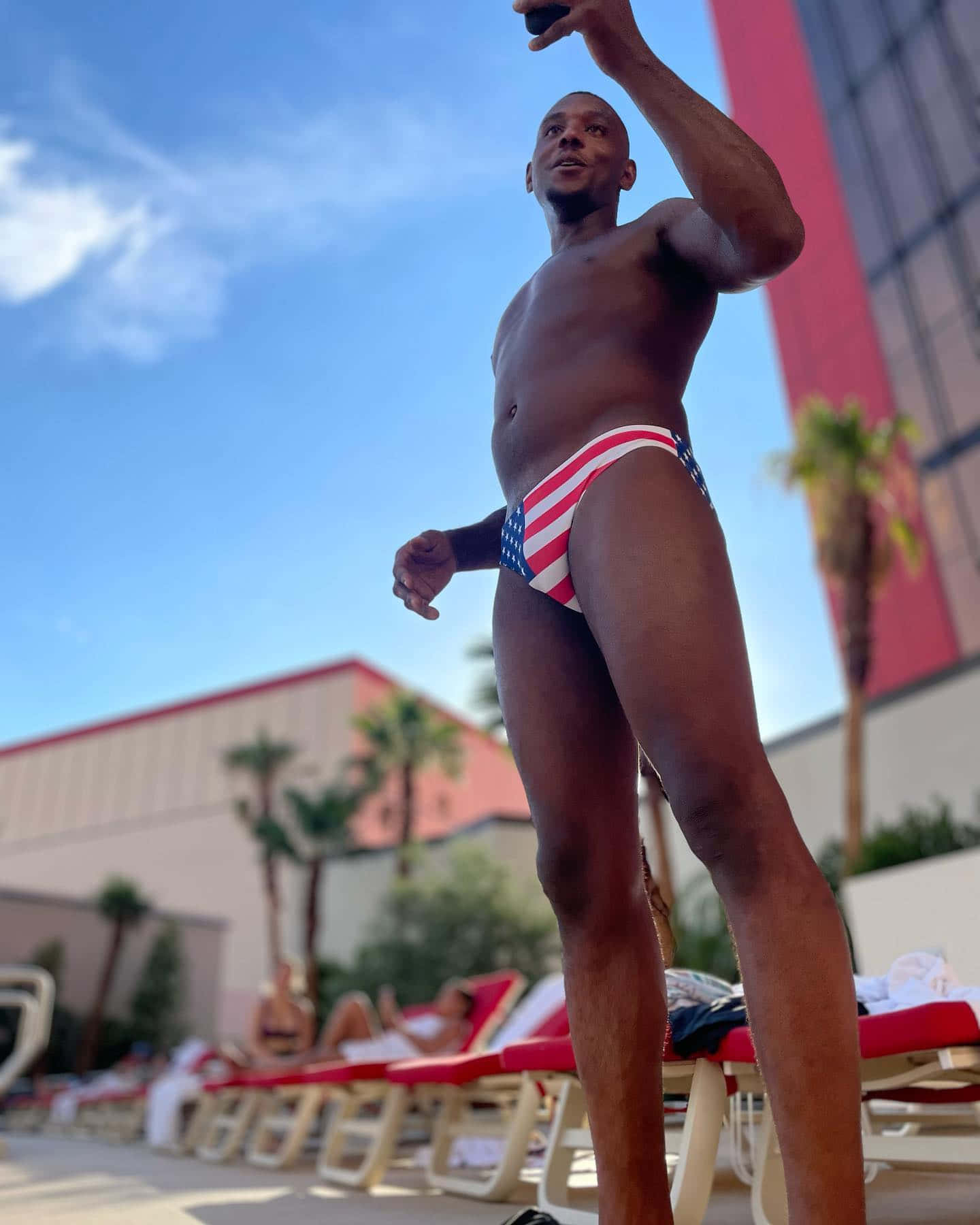 Vacation 桌布 壁紙 — 一位肌肉發達的男子穿著愛國泳褲，在陽光普照的度假村泳池旁，棕櫚樹和躺椅環繞下自拍。