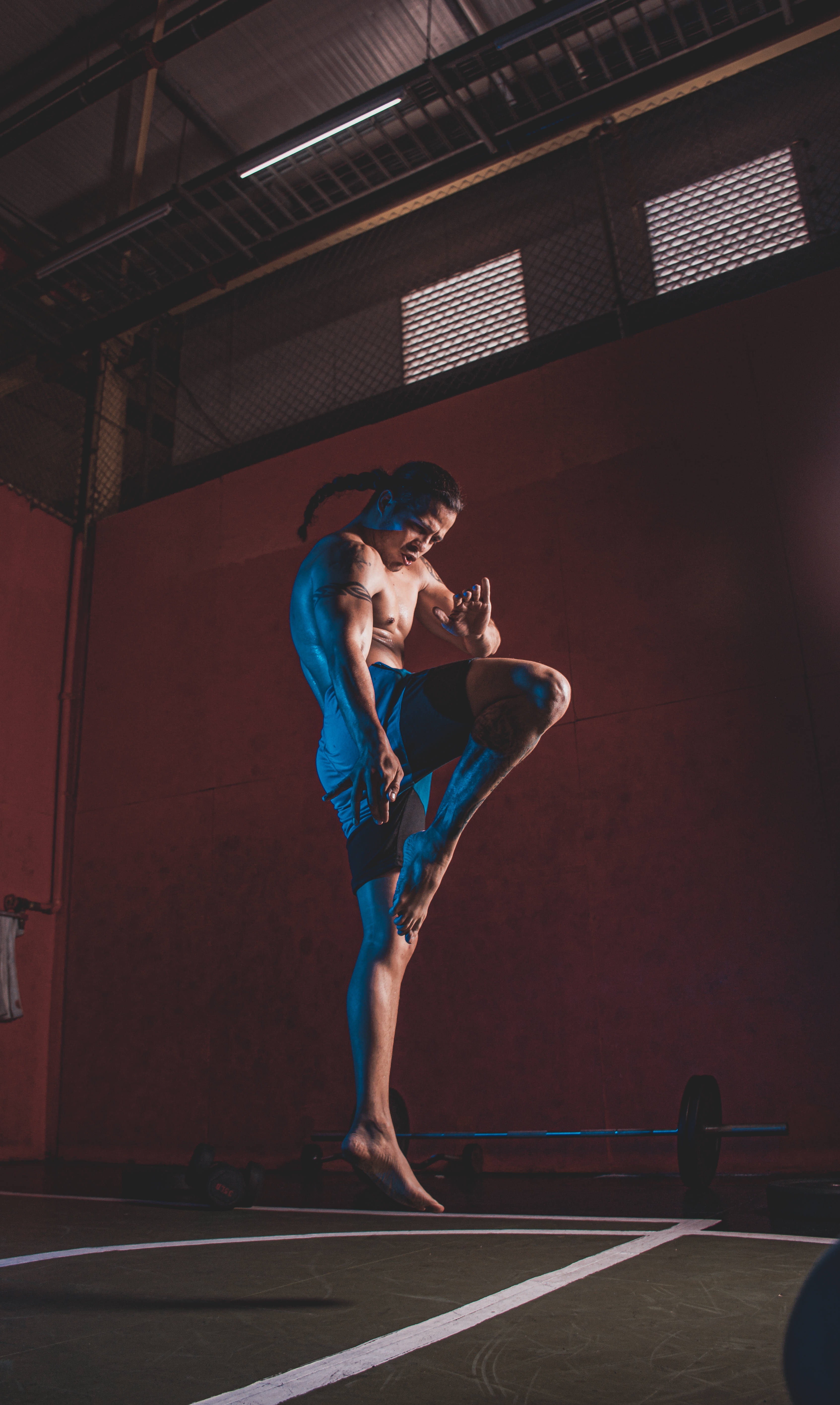 MMA 桌布 壁紙 — 一位肌肉發達的男子穿著藍色短褲，在光線昏暗、紅牆和裸露天花橫樑的健身房中執行高踢腿動作。