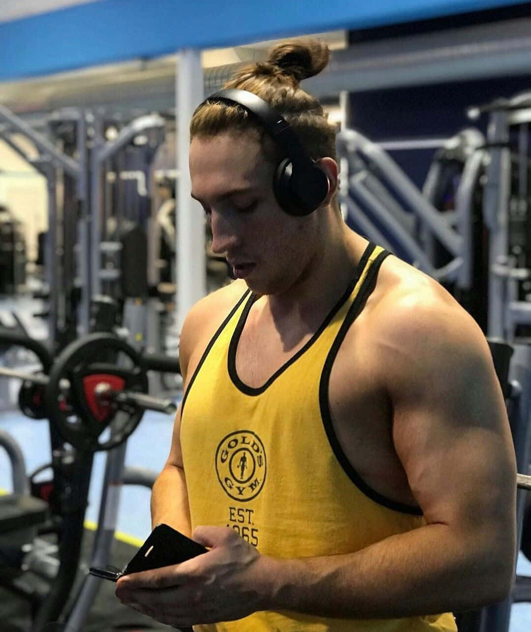 Workout 桌布 壁紙 — 一位肌肉發達的男子身穿黃色Gold's Gym背心，戴著耳機，在健身器材中查看手機。