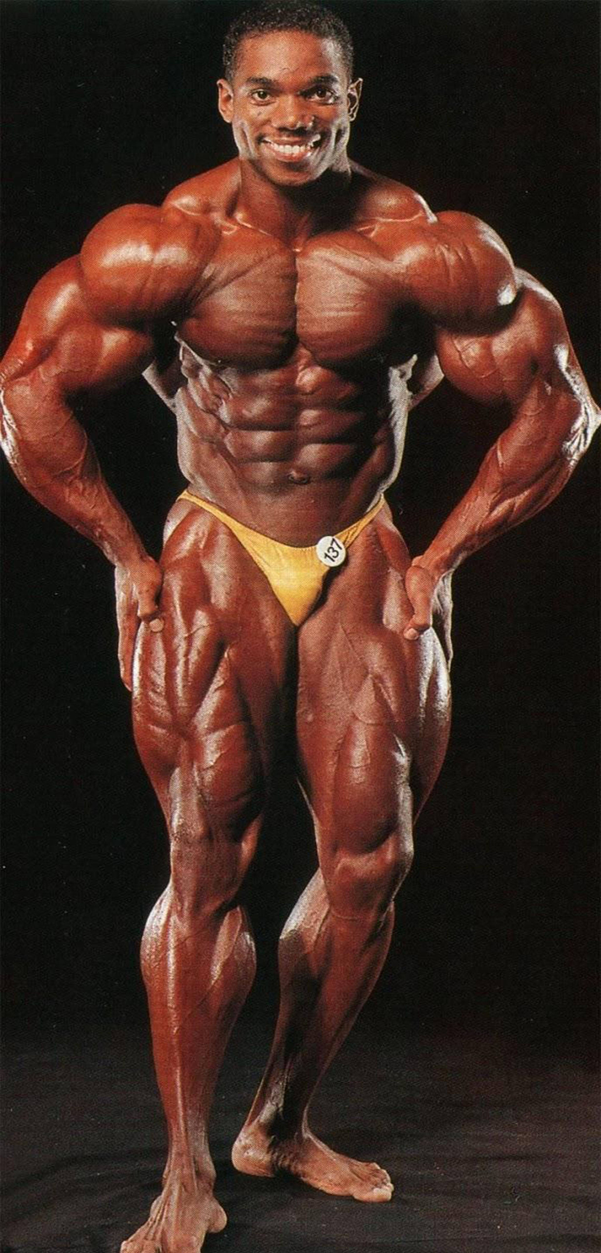 flex wheeler 桌布 壁紙 — 一位肌肉發達的男性健美運動員自信地穿著黃色比賽褲在黑色背景前擺姿勢。