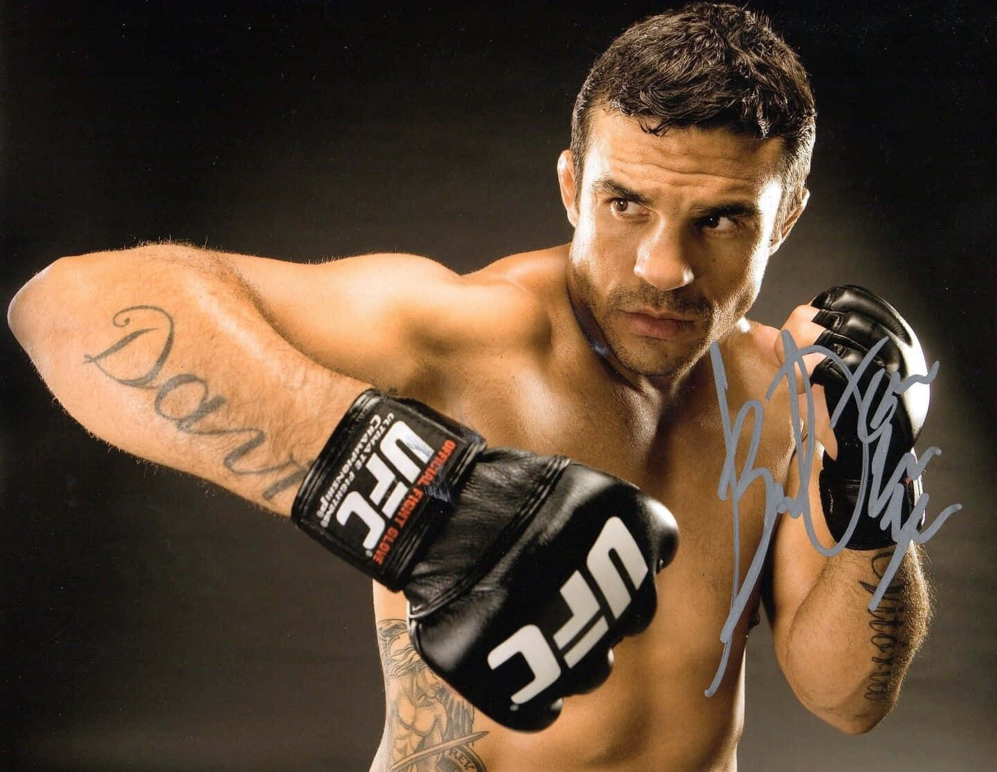 UFC 桌布 壁紙 — 一位肌肉發達的男性拳手戴著黑色UFC手套，在黑暗的攝影棚背景下擺出強有力的姿勢，身上有可見的紋身和簽名。