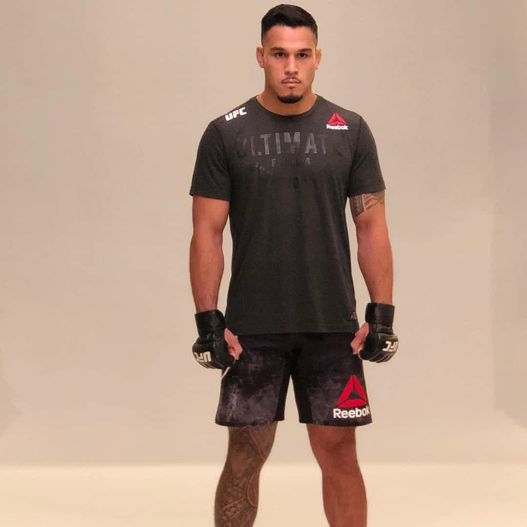 UFC 桌布 壁紙 — 一位肌肉發達的男性拳手身穿UFC裝備，戴著手套和刺青，自信地站在純色工作室背景前。
