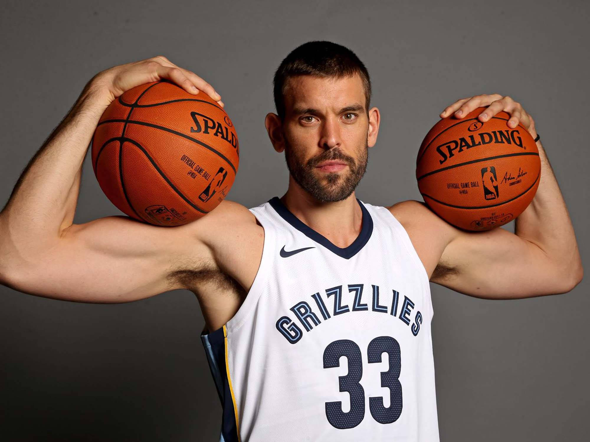 Marc Gasol 桌布 壁紙 — 一位肌肉發達的籃球運動員身穿灰熊隊球衣，在灰色工作室背景下將兩個 Spalding 籃球扛在肩上。