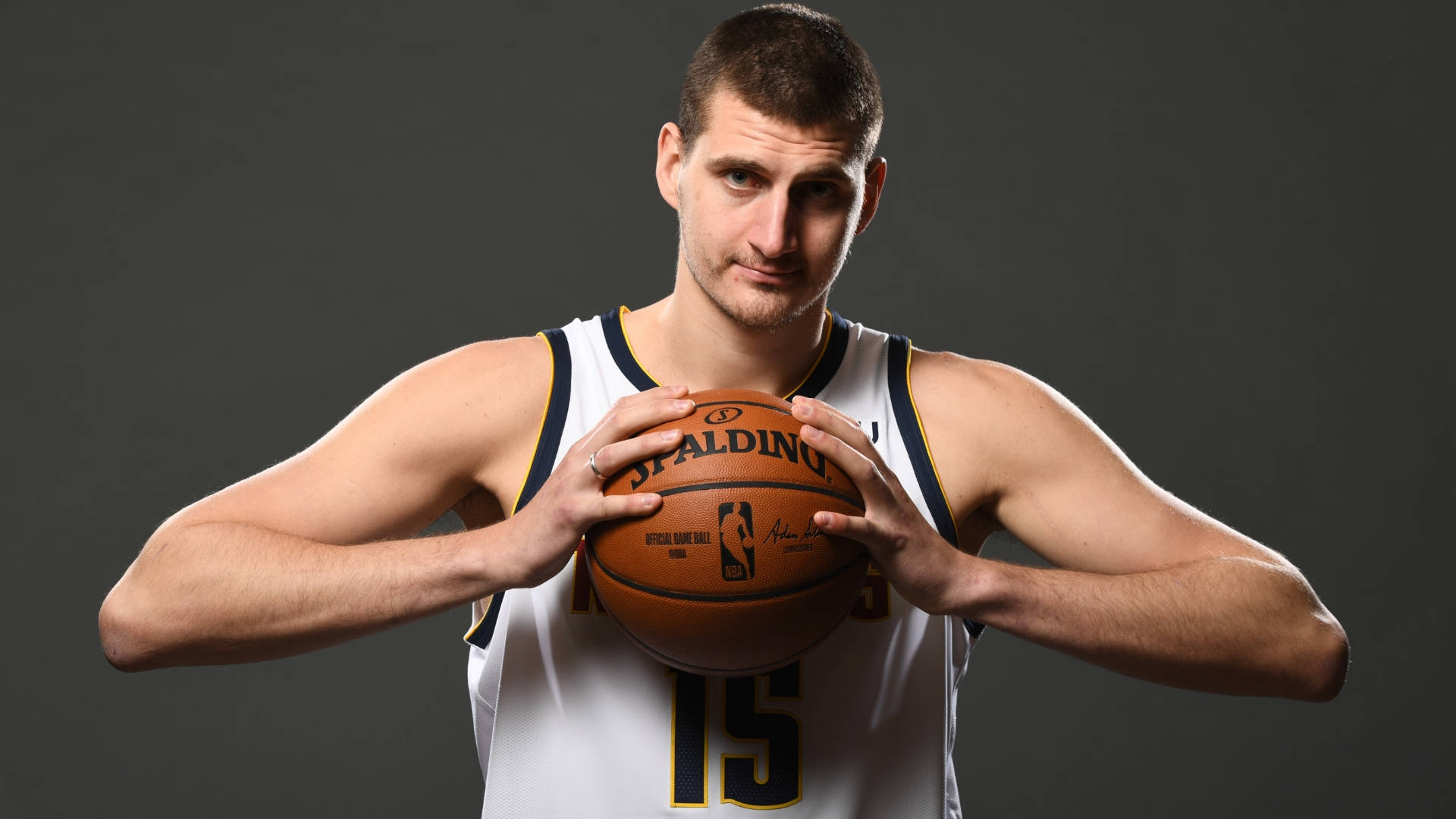 Jokic 桌布 壁紙 — 一位肌肉發達的籃球運動員身穿白色球衣，在黑暗的攝影棚背景下，專注地拿著一個 Spalding 籃球。
