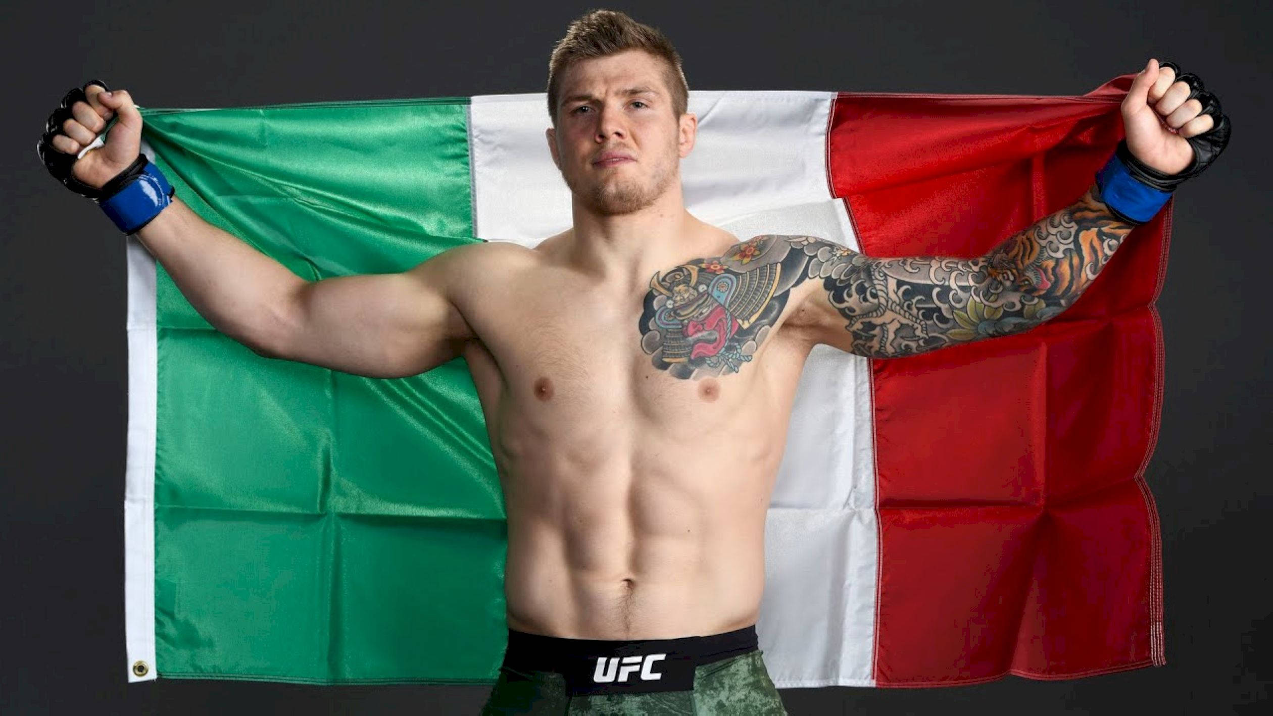 UFC 桌布 壁紙 — 一位肌肉發達的義大利MMA拳手，在黑暗的攝影棚背景下，用刺青的手臂自豪地高舉國旗。