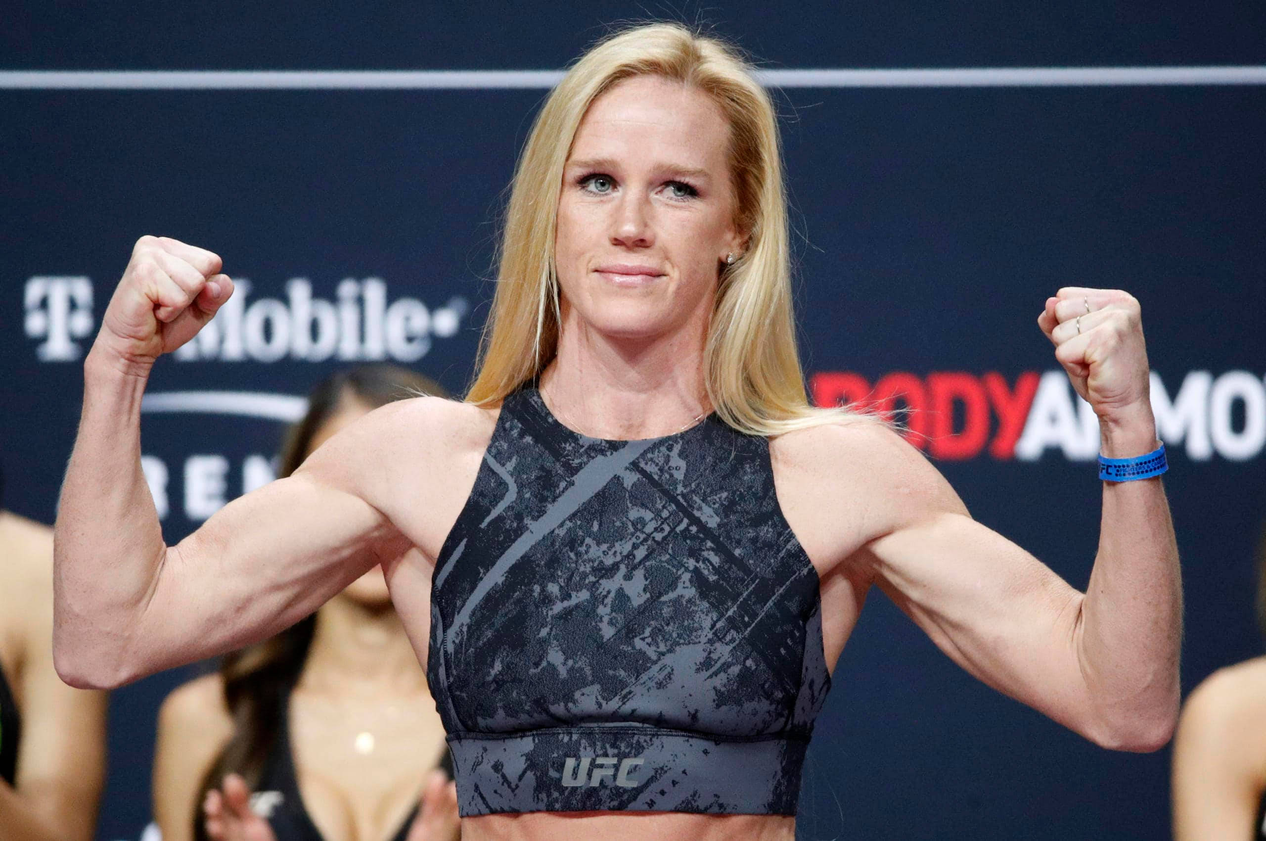 MMA 桌布 壁紙 — 一位肌肉發達的金髮女性在品牌背景的稱重活動中，穿著UFC運動內衣彎曲雙臂。