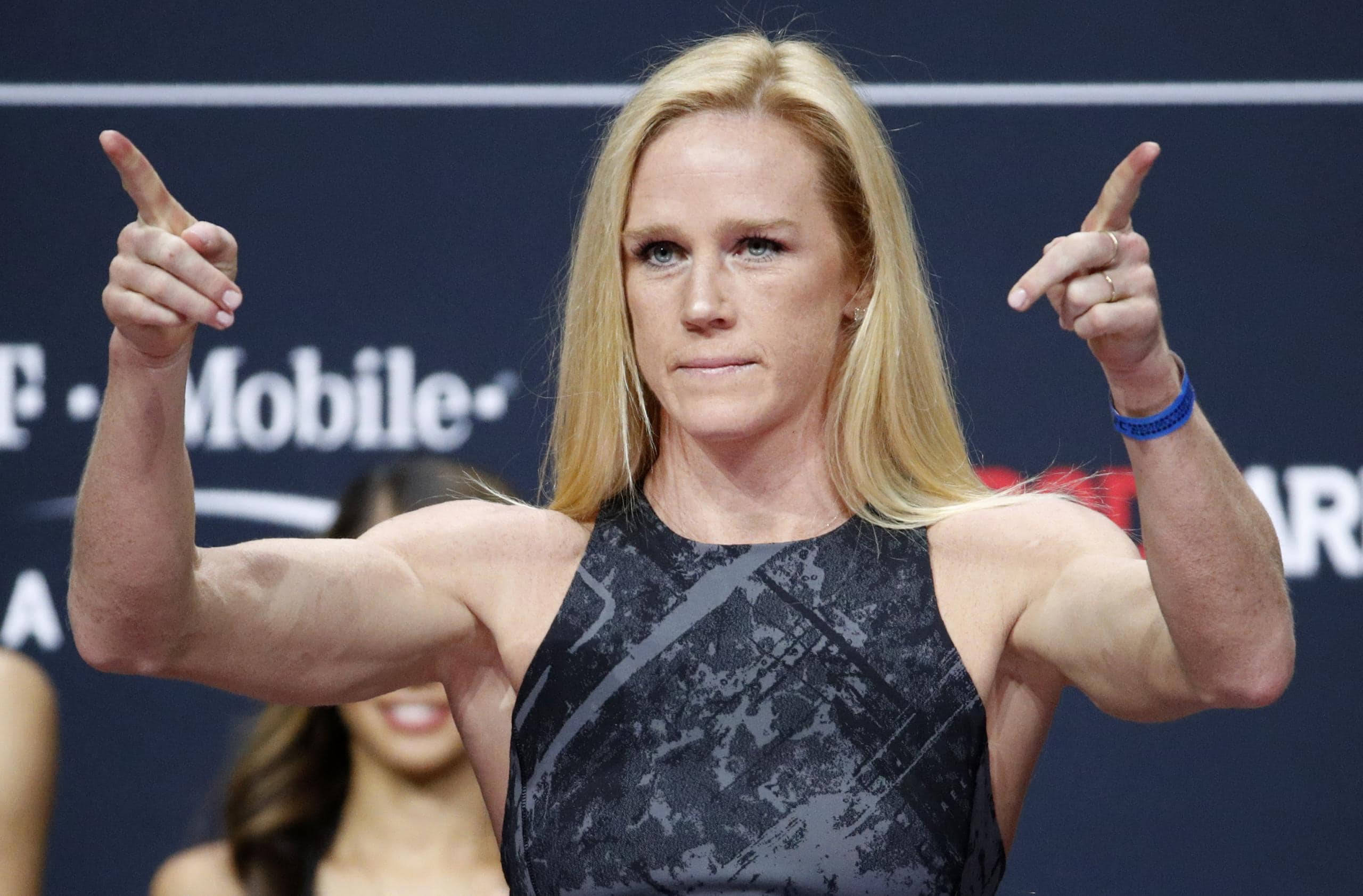 MMA 桌布 壁紙 — 一位肌肉發達的金髮女性穿著黑色圖案上衣，在藍色品牌背景前，專注地向前伸出雙食指。