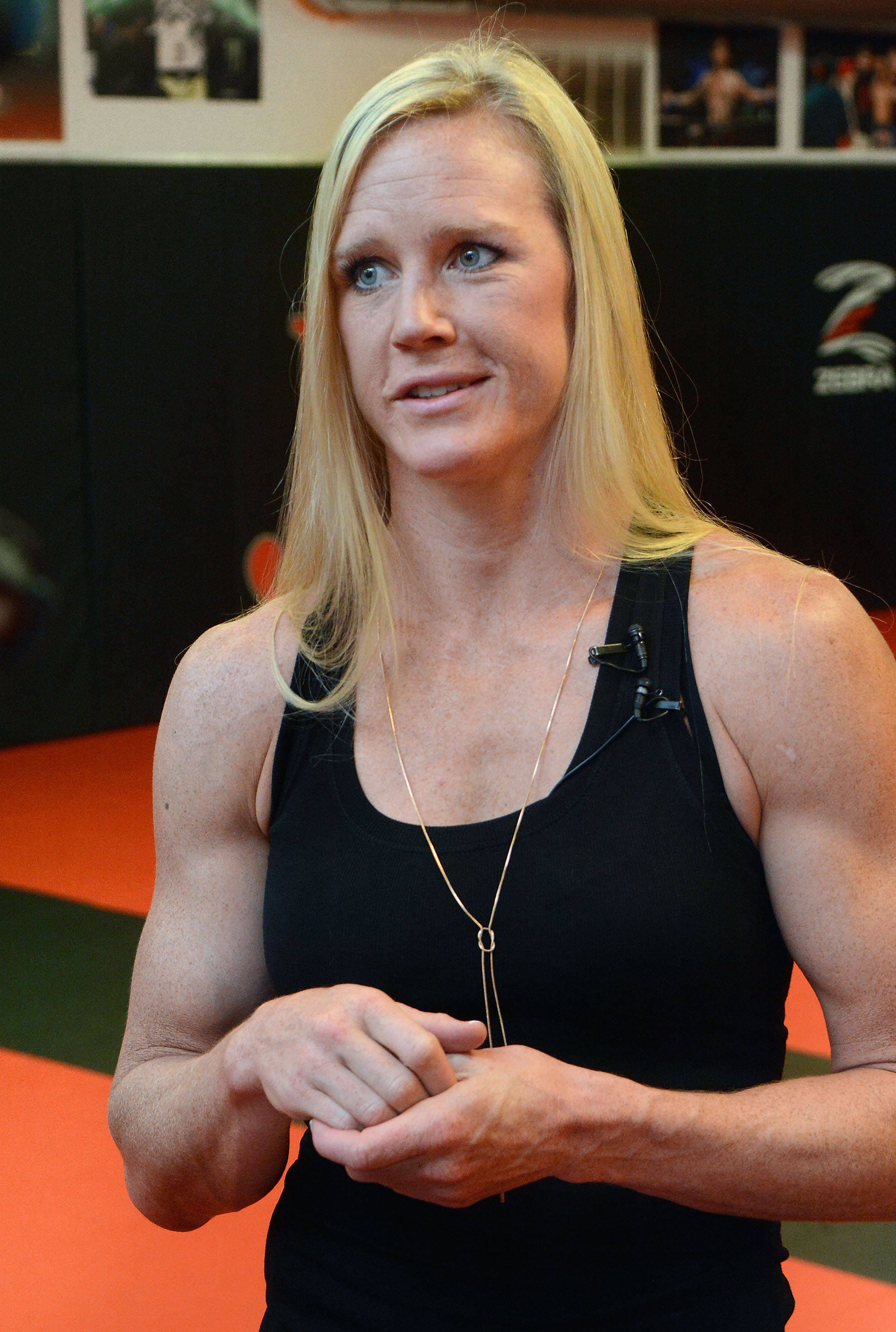Strong Woman 桌布 壁紙 — 一位肌肉發達的金髮女性穿著黑色背心，自信地站在一個有橙色墊子和拳擊海報的健身房裡。