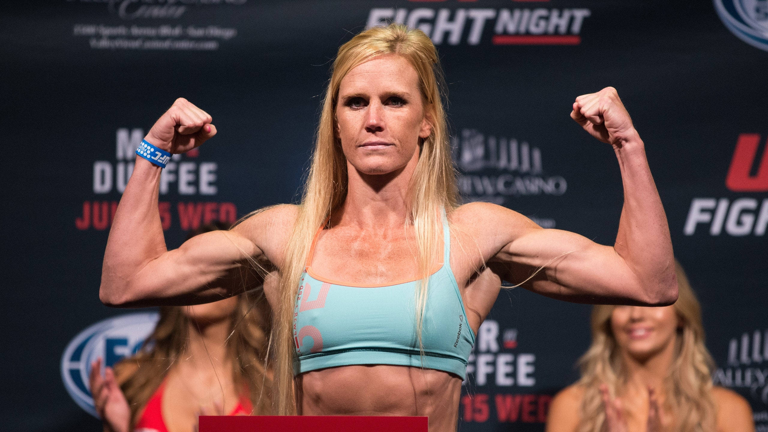MMA 桌布 壁紙 — 一位肌肉發達的金髮女拳手在稱重活動中彎曲雙臂，身後有「FIGHT NIGHT」品牌標誌。