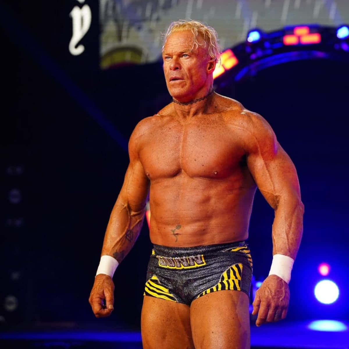 WWE 桌布 壁紙 — 一位肌肉發達的金髮摔角手，穿著黑色和黃色緊身褲，在藍色和紅色燈光下的黑暗舞台上被強烈照亮。
