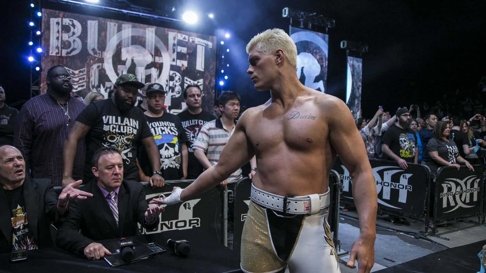 WWE 桌布 壁紙 — 一位肌肉發達的金髮摔角手穿著金色和白色的短褲，自信地站在擂台上，手持冠軍腰帶，粉絲和官員們專注地觀看。