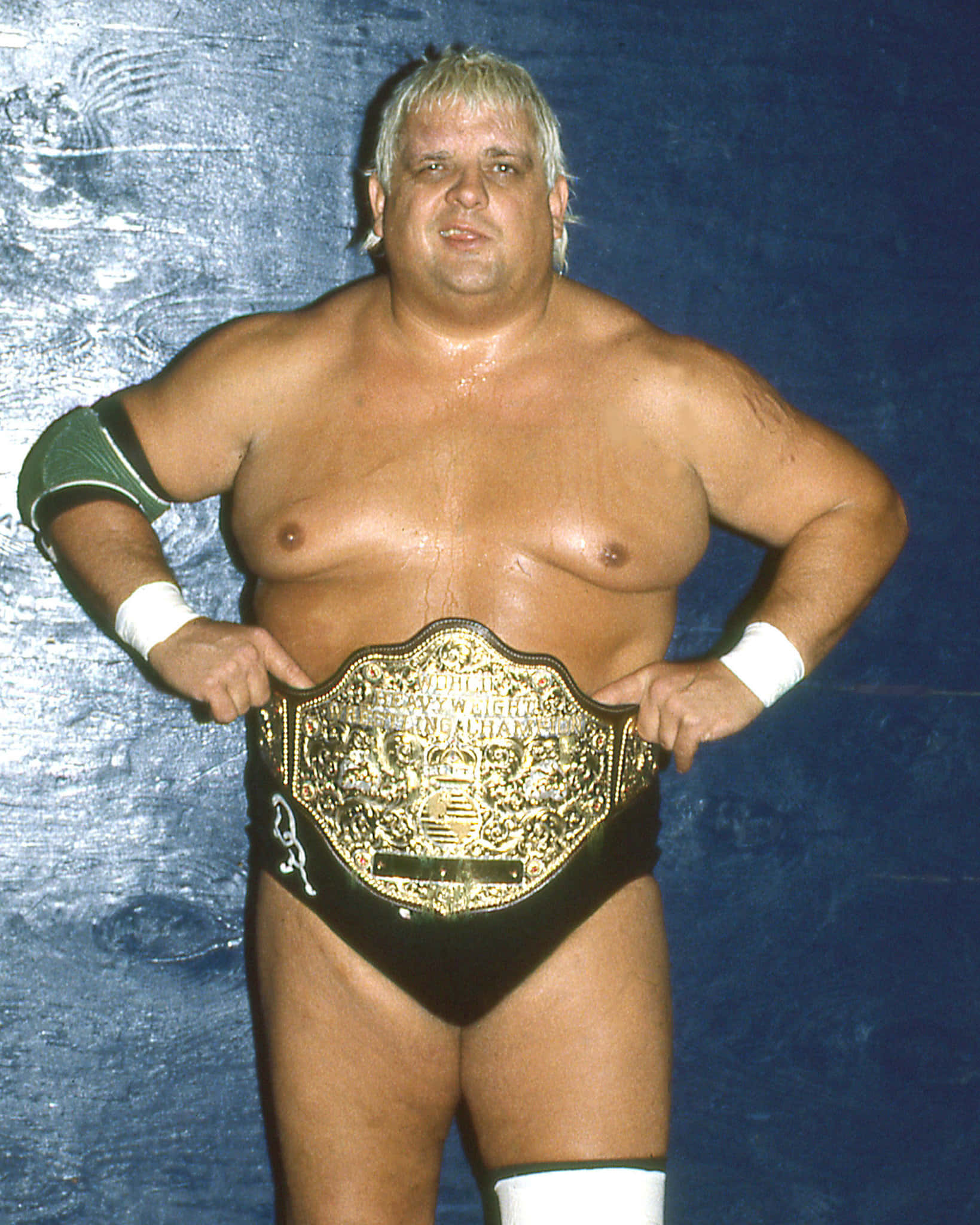 Dusty Rhodes 桌布 壁紙 — 一位肌肉發達的金髮摔角手自信地站在有紋理的藍色背景前，手持金色冠軍腰帶。