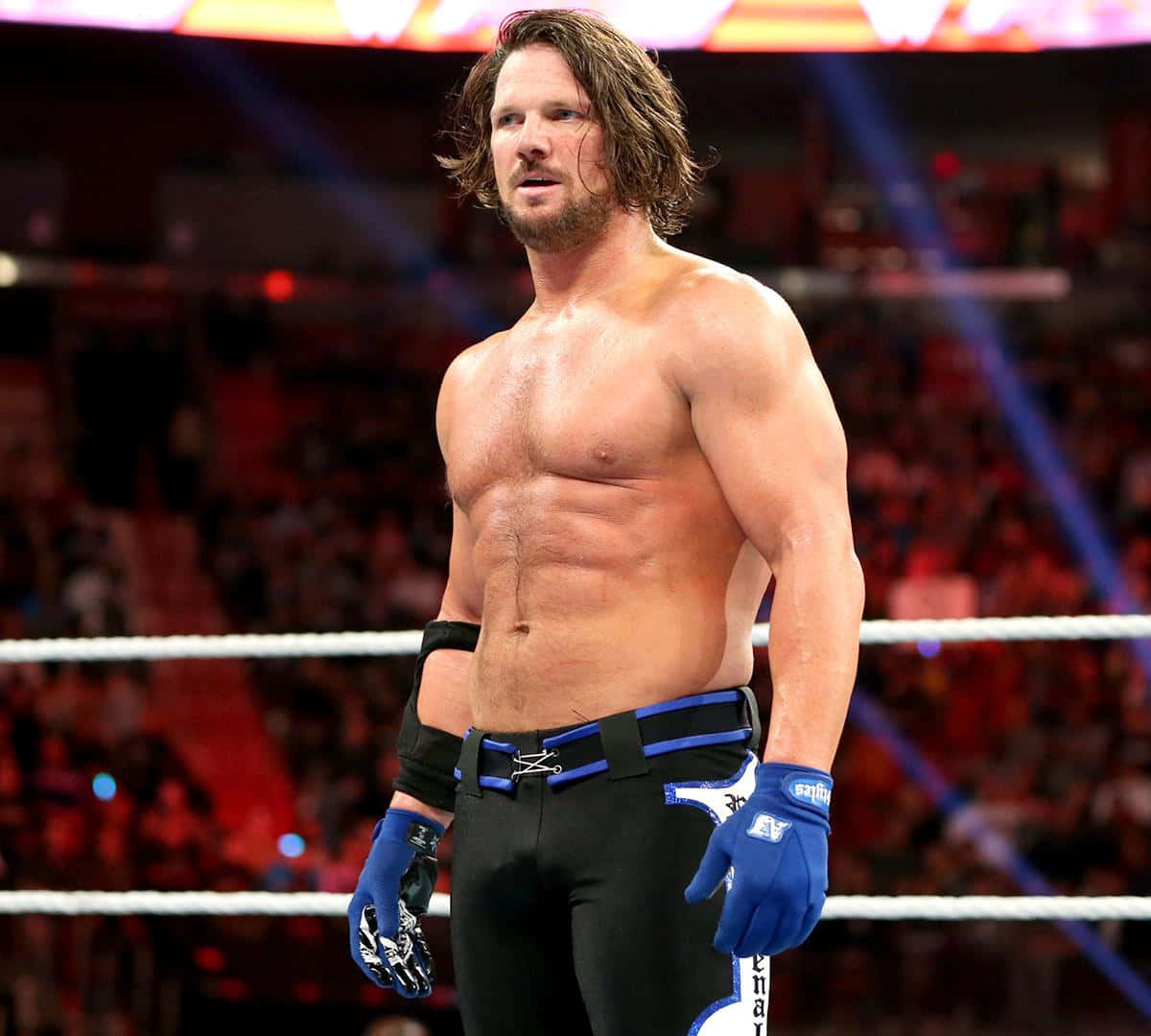 WWE 桌布 壁紙 — 一位肌肉發達的長髮摔角手站在擂台上，穿著黑色短褲和藍色手套，在明亮的場館燈光下。