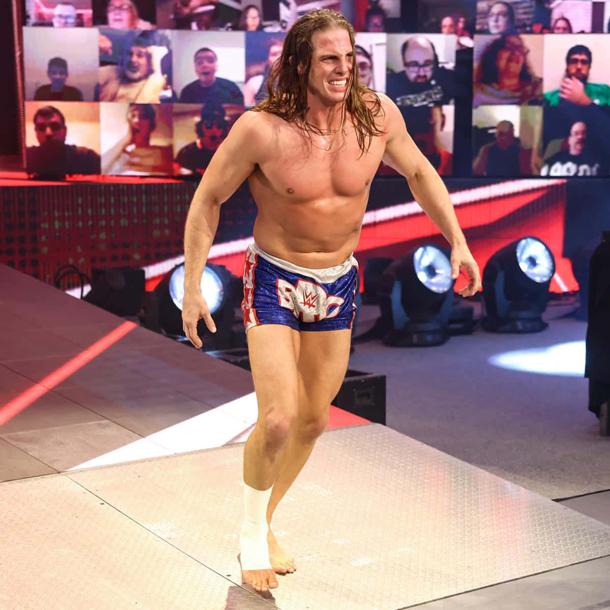 WWE 桌布 壁紙 — 一位肌肉發達的長髮摔角選手，在明亮的舞台燈光下，穿著藍、紅、白三色短褲，自信地走下斜坡。