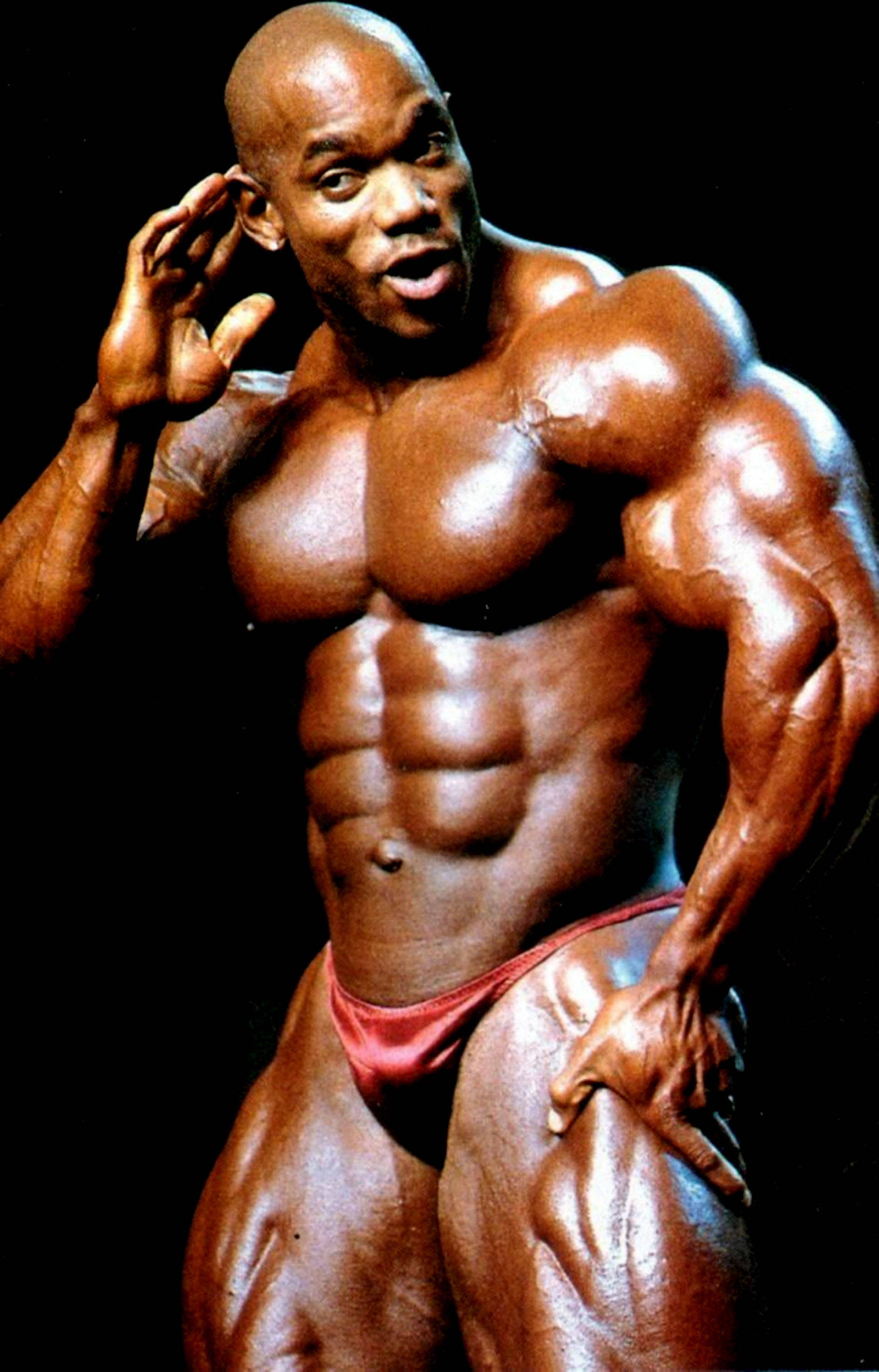 flex wheeler 桌布 壁紙 — 一位肌肉發達的黑人健美運動員在黑色背景前穿著紅色比賽褲，展示清晰的腹肌和手臂。