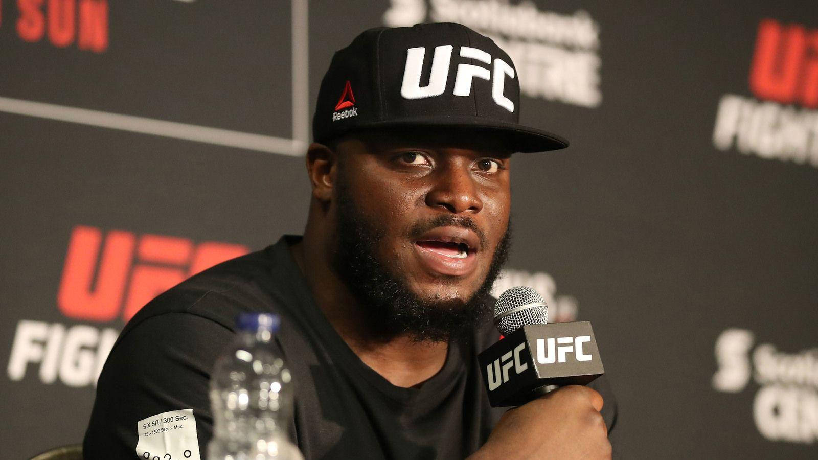 UFC 桌布 壁紙 — 一位肌肉發達的黑人拳手戴著UFC帽子，在有品牌背景的新聞發布會上對著麥克風講話。