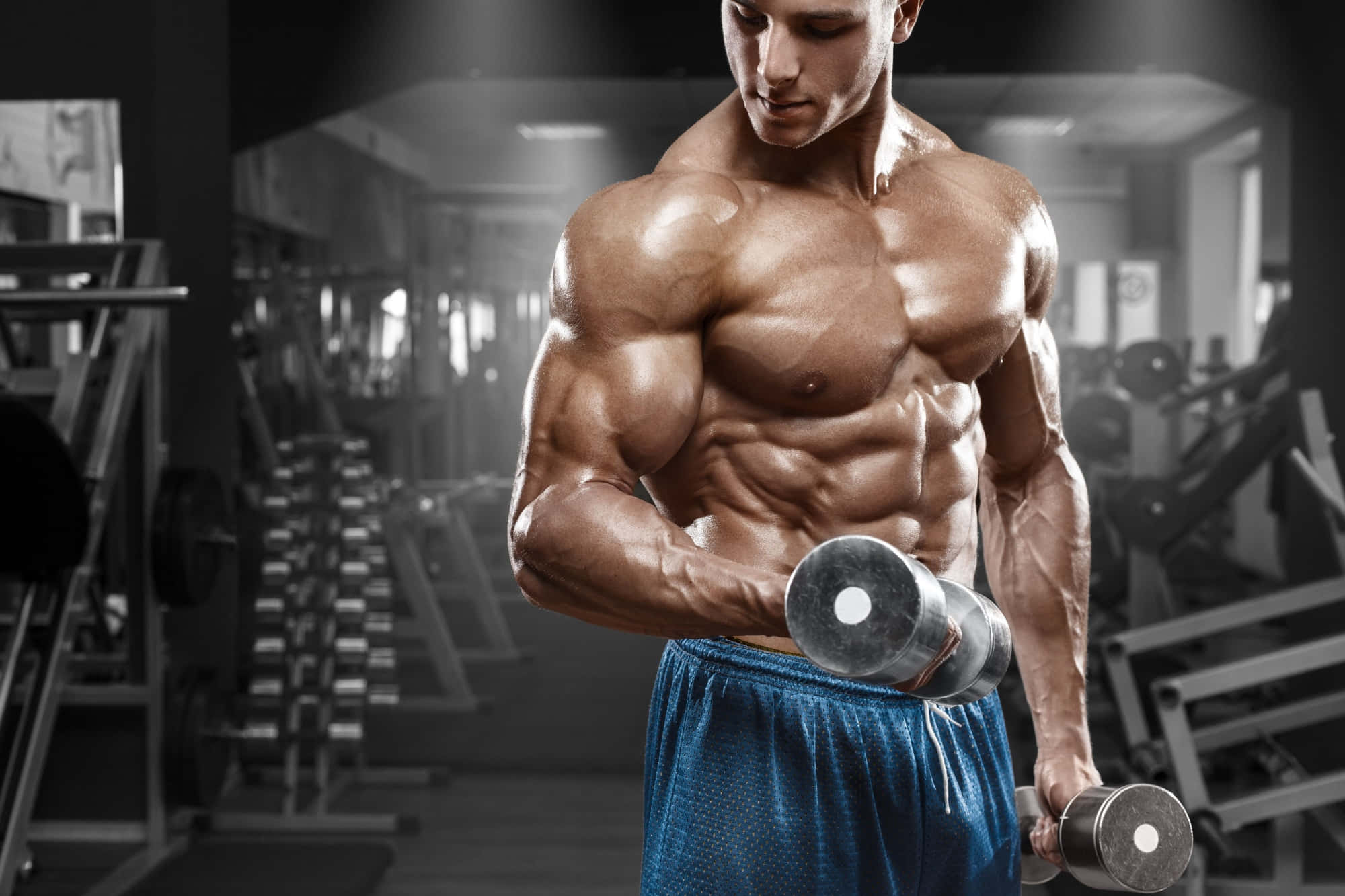 Muscle 桌布 壁紙 — 一位肌肉線條分明、腹肌明顯的男子在燈光昏暗的健身房中舉起啞鈴，背景中的器材模糊不清。
