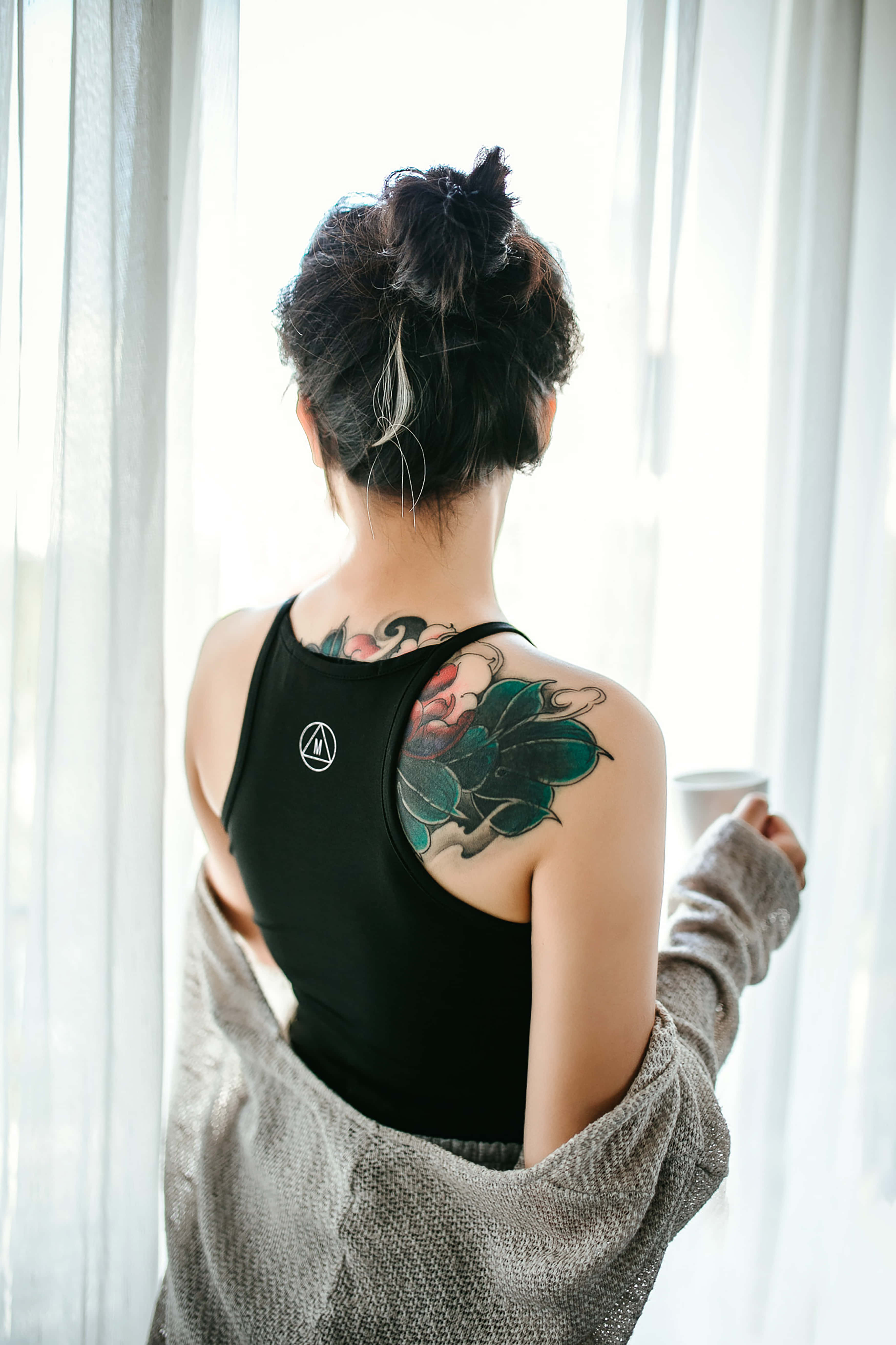 Tattoo Girl 桌布 壁紙 — 一位肩上帶有花卉刺青的女士，穿著黑色背心和灰色毛衣站在窗邊。
