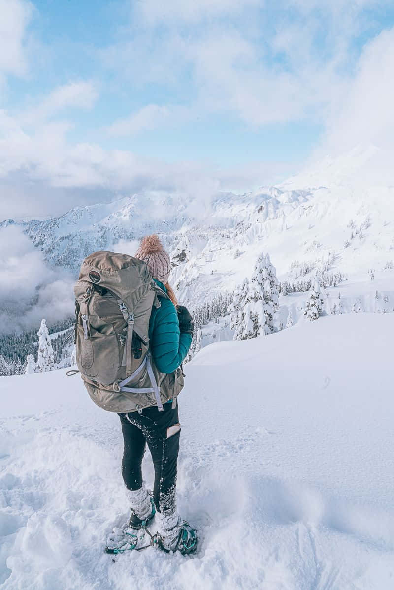Winter Hiking 桌布 壁紙 — 一位背著大背包的遠足者站在白雪覆蓋的山頂上，俯瞰著部分多雲天空下廣闊的冬季景觀。