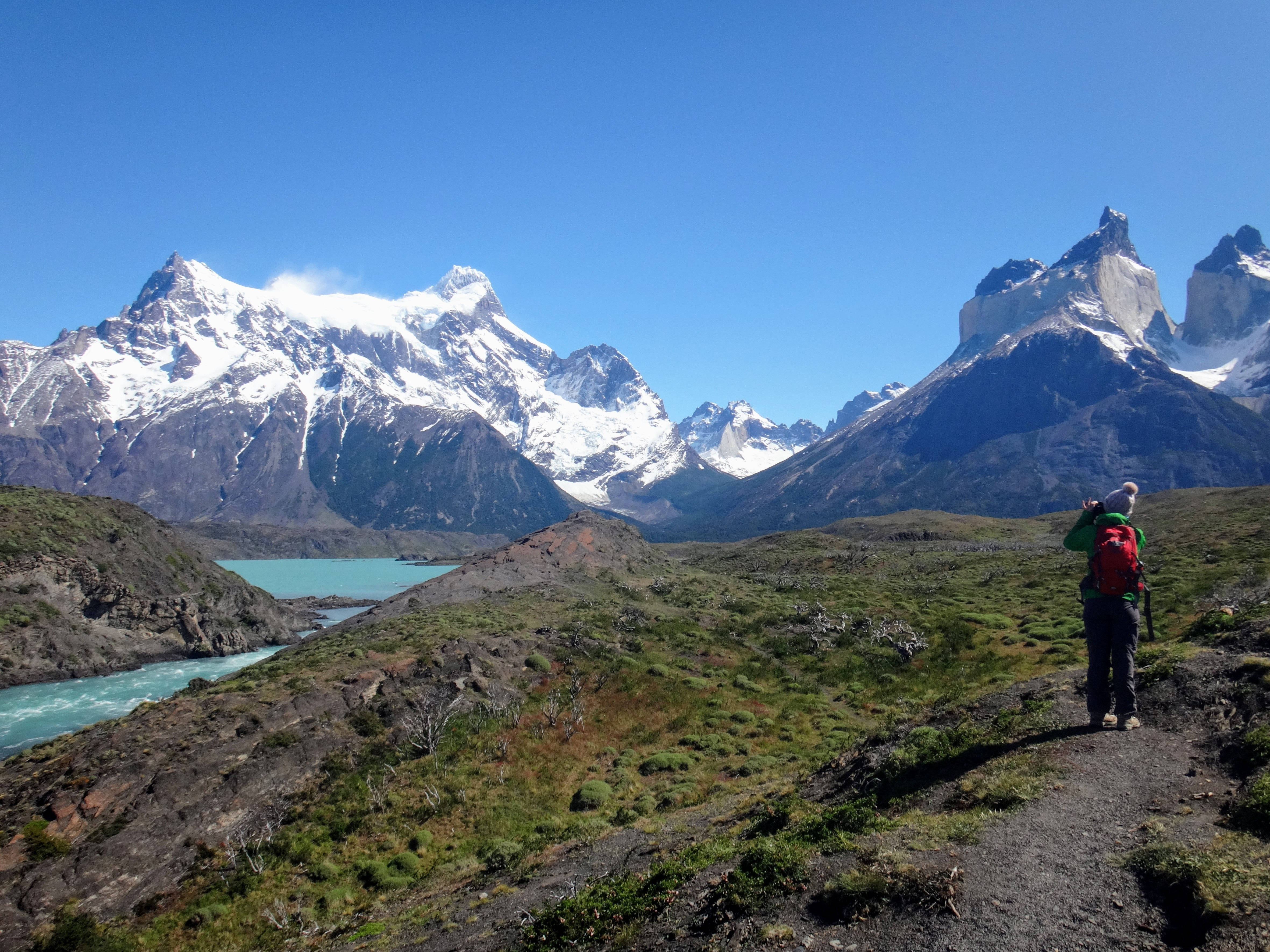 Motivation 桌布 壁紙 — 一位背著紅色背包的徒步旅行者站在小徑上，凝視著白雪覆蓋的山峰和碧綠的湖泊，天空湛藍清澈。