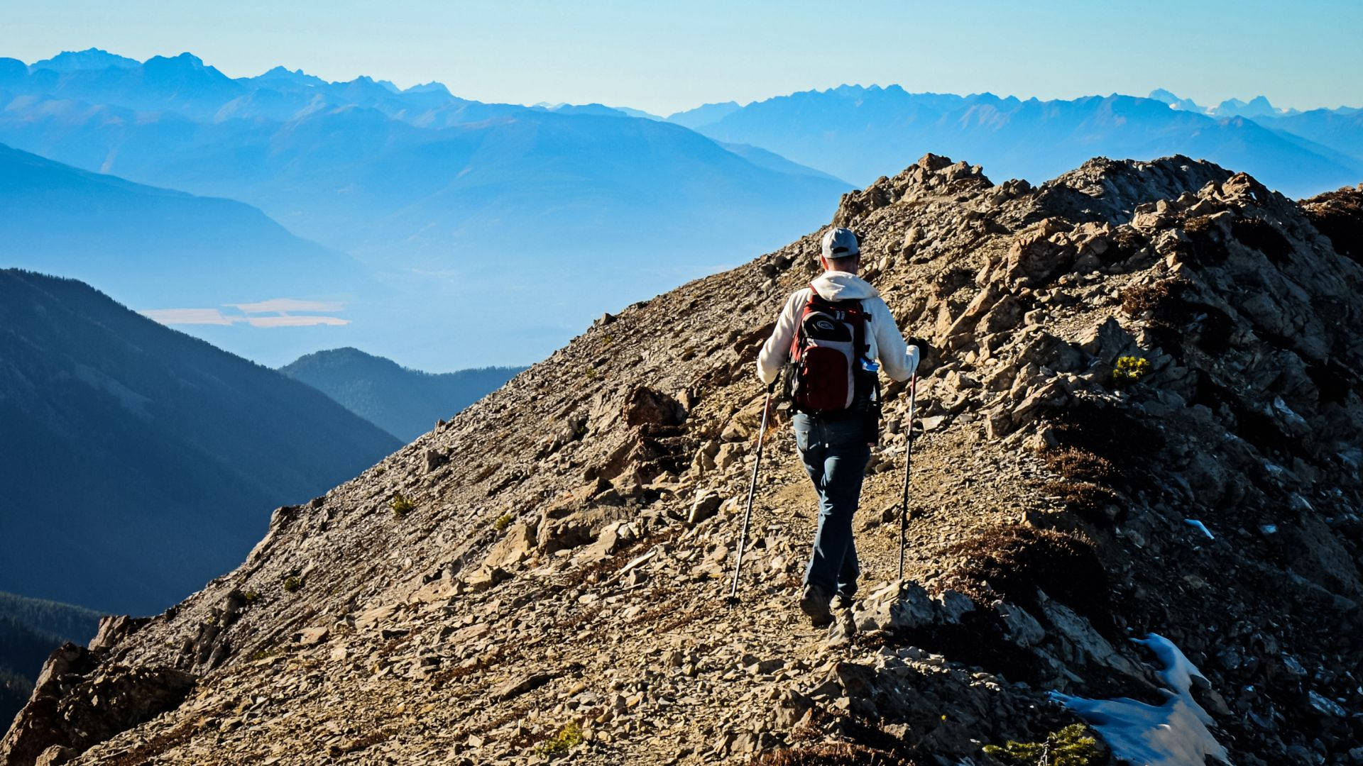 Man 桌布 壁紙 — 一位背著背包、拿著登山杖的健行者，在晴朗藍天下攀爬岩石山脊，下方遠處可見山峰和山谷。
