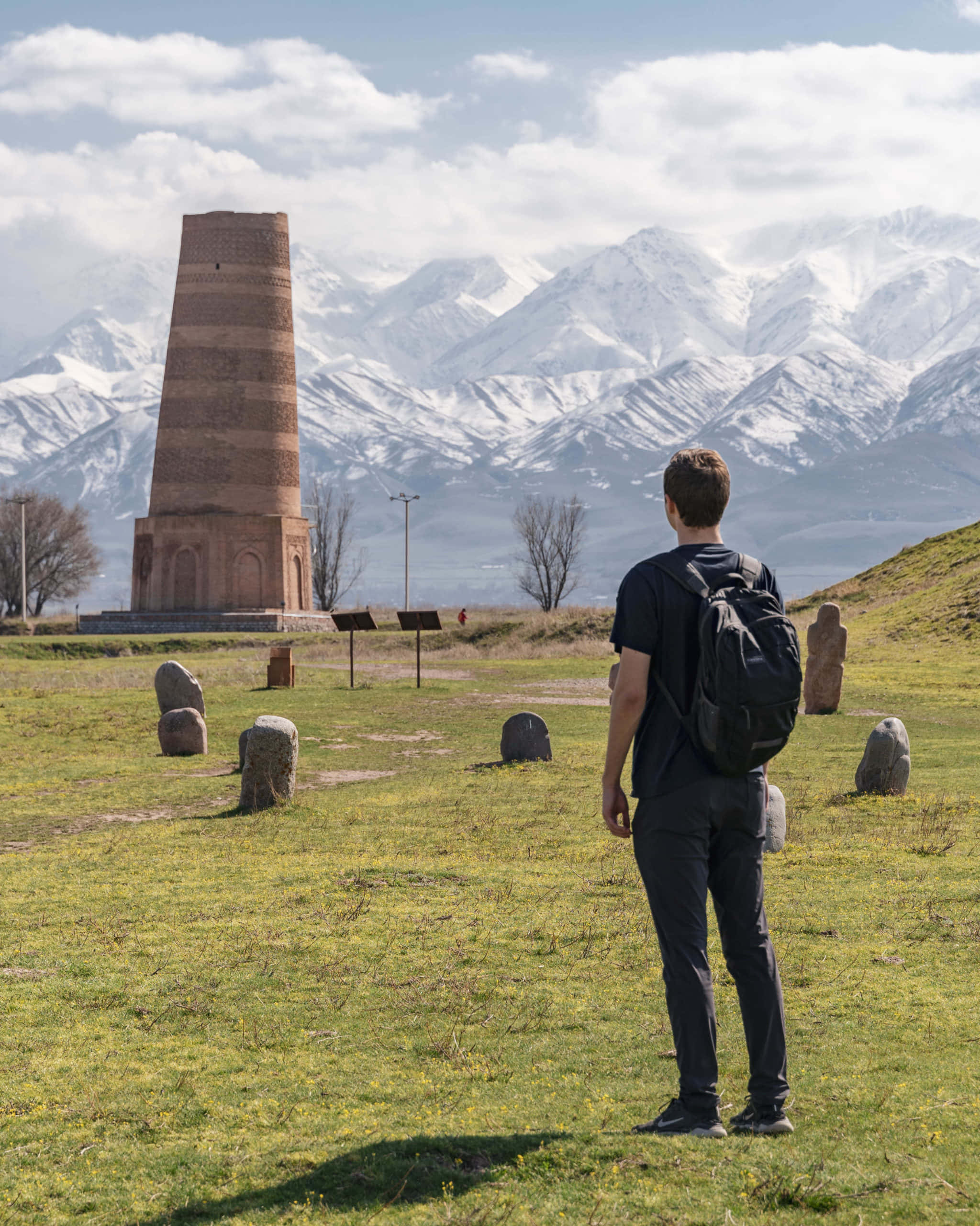 Kyrgyzstan 桌布 壁紙 — 一位背著背包的旅行者站在草地上，凝視著一座古老的磚塔，背後是白雪覆蓋的山峰。