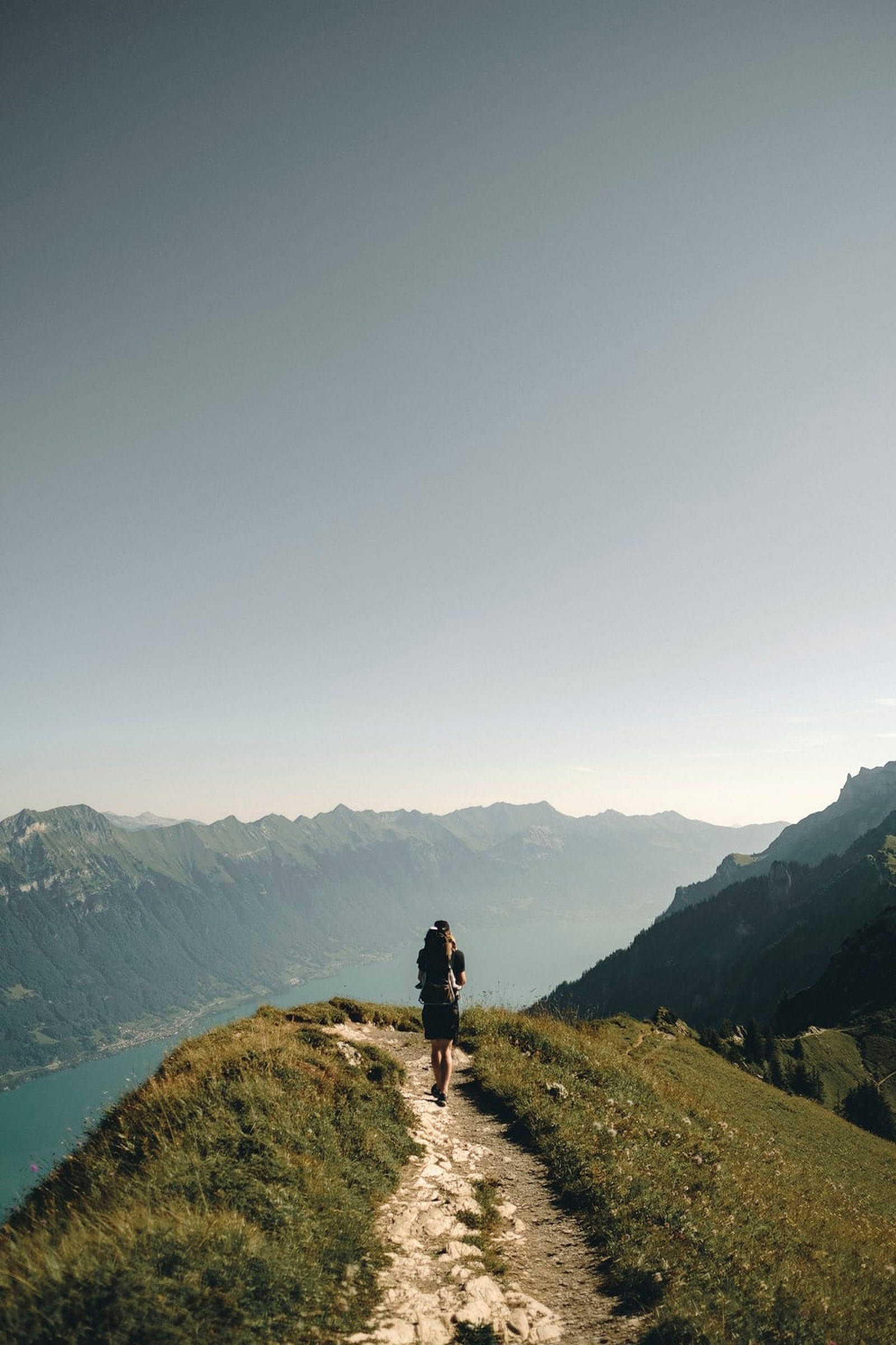登山 桌布 壁紙 — 一位背著背包的獨行登山者，沿著狹窄的山脊步道行走，俯瞰廣闊的山谷和碧綠的湖泊，天空晴朗。